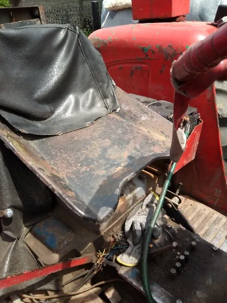 Zbliżenie na fragment starego, czerwonego traktora. Widoczne jest czarne, skórzane lub gumowe siedzisko z przetarciami, zardzewiałe metalowe elementy nadwozia oraz fragmenty okablowania i zielony wąż w dolnej części obrazu.