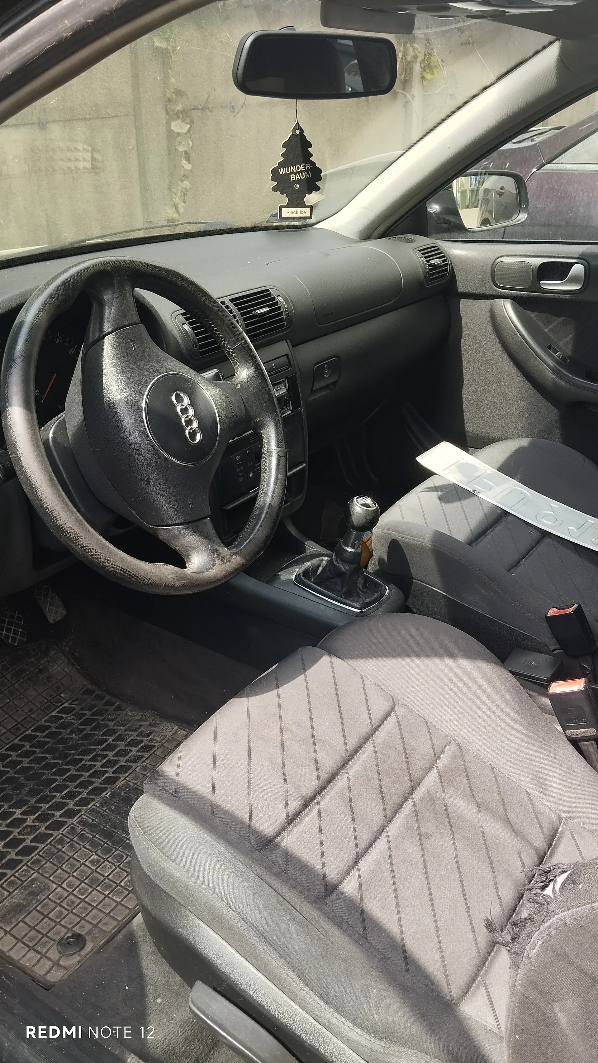 Wnętrze samochodu marki Audi z czarną deską rozdzielczą, kierownicą z logo Audi, manualną skrzynią biegów i szarymi siedzeniami z pionowymi prążkami. Na lusterku wstecznym wiszący aromatyzator powietrza w kształcie choinki z napisem "WUNDER-BAUM" oraz "Black Ice". Na siedzeniu pasażera widoczna tablica rejestracyjna. W tle przez przednią szybę betonowa ściana i fragment innego pojazdu. W lewym dolnym rogu napis "REDMI NOTE 12".