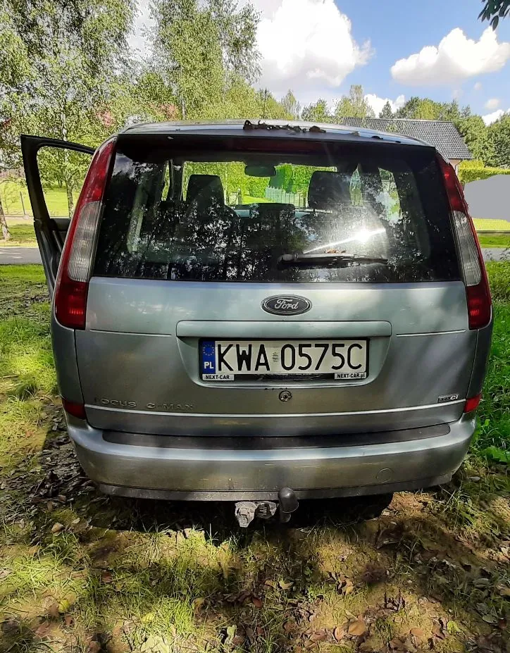Tylny widok srebrnego samochodu marki Ford Focus C-Max zaparkowanego na trawie. Pojazd ma polską tablicę rejestracyjną o numerze KWA 0575C. Widoczny jest hak holowniczy oraz otwarte lewe tylne drzwi. W tle znajdują się zielone drzewa i niebo z białymi chmurami.