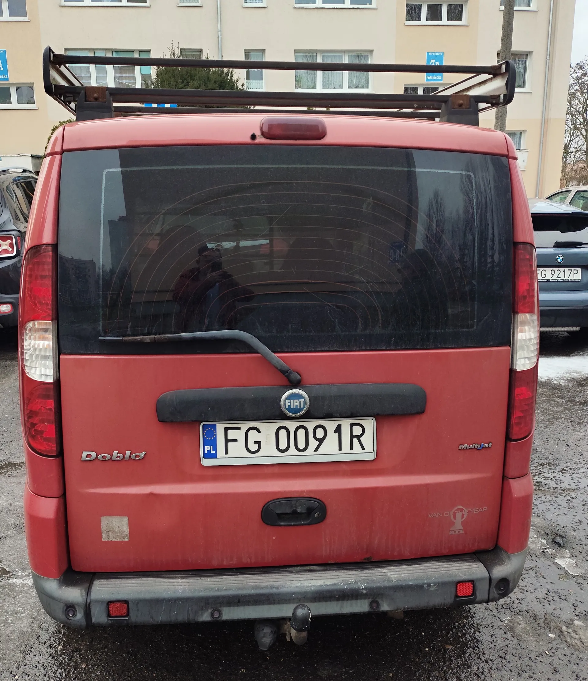 Czerwony samochód dostawczy Fiat Doblo z tyłu, z widocznym tablicą rejestracyjną FG 0091R (PL), napisem Doblo po lewej stronie, logo Fiat w centrum i napisem Multijet po prawej. Na dachu znajduje się czarny bagażnik dachowy. W tle budynek mieszkalny o beżowej elewacji z oknami oraz częściowo widoczne inne pojazdy, w tym samochód z tablicą G 9217P. Podłoga parkingowa jest mokra, a na tylnej szybie widoczne są refleksy drzew i budynków.