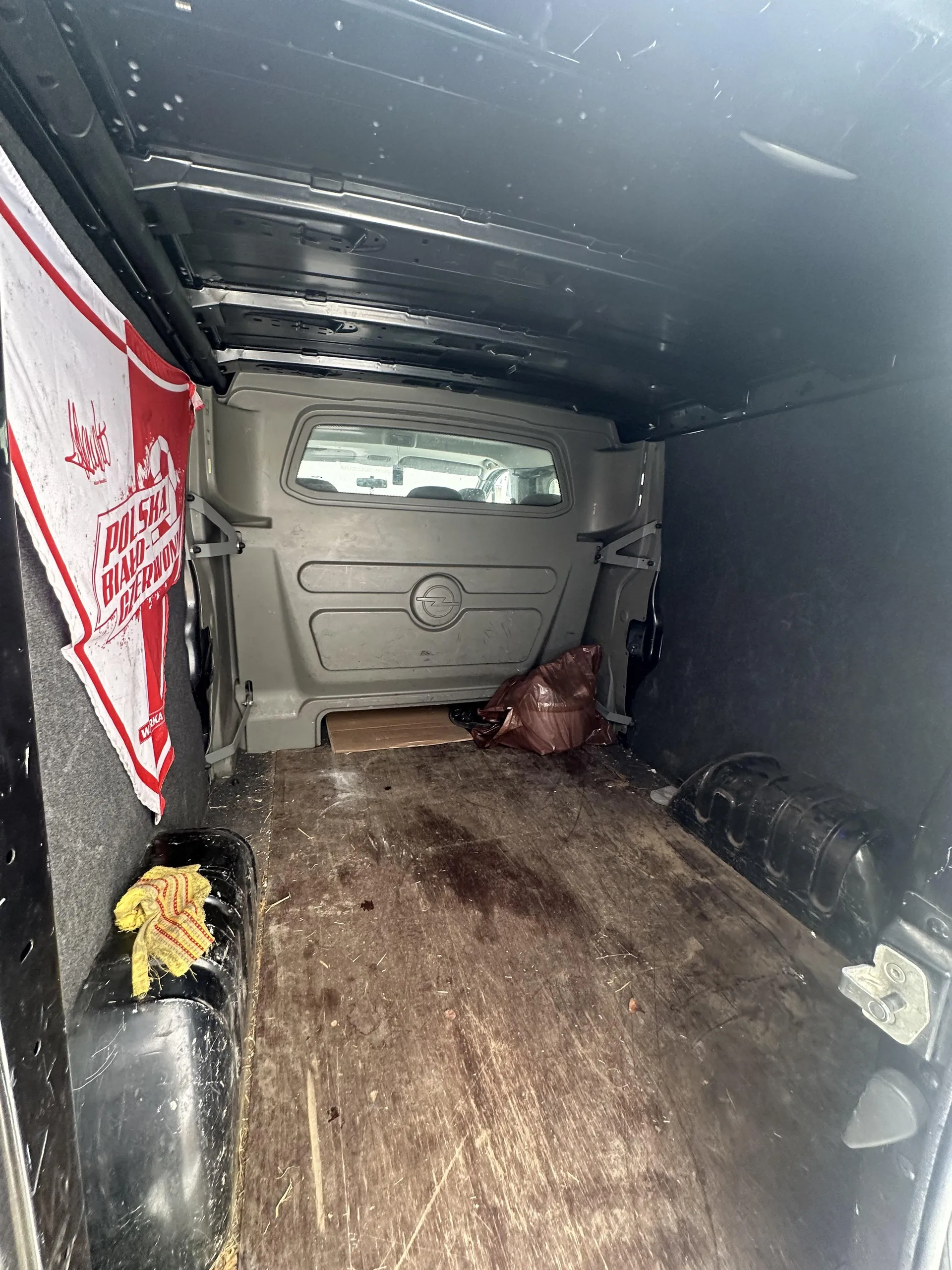 Wnętrze furgonetki z widoczną tylną częścią kabiny kierowcy. Po lewej stronie wisząca czerwono-biała flaga z napisem "Polska Biało-Czerwona" oraz mniejszym napisem "Warka" na dole. Na podłodze z brudnymi plamami leży brązowa torba i kawałek kartonu. W dolnej części po lewej stronie czarny przedmiot z żółtą tkaniną w czerwone paski. Ściany i sufit furgonetki są ciemnoszare, podłoga wydaje się być z drewna lub podobnego materiału.