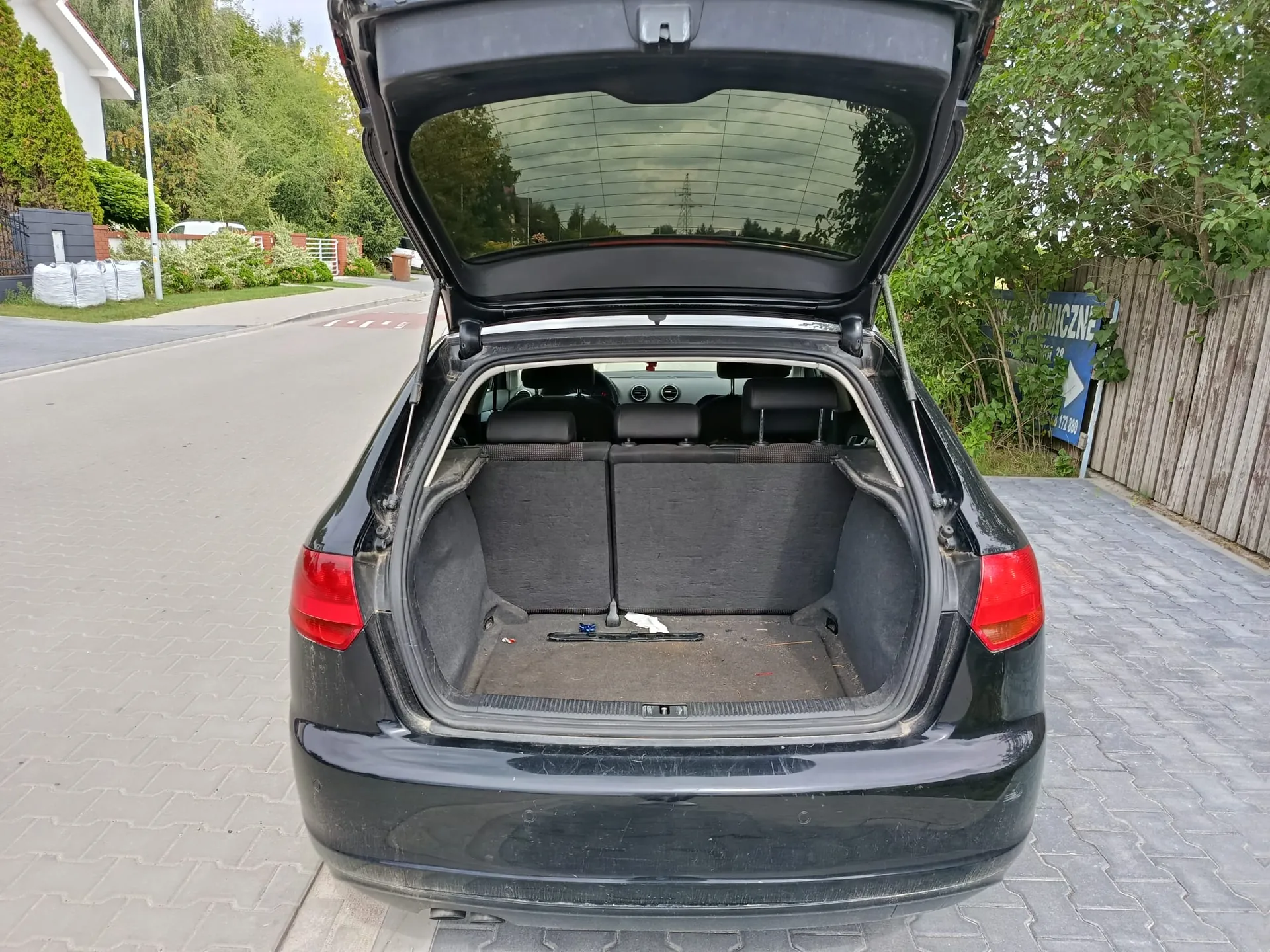 Tylny widok czarnego samochodu z otwartym bagażnikiem. Wnętrze bagażnika jest wyłożone szarym materiałem i jest niemal puste. W tle widoczna jest utwardzona droga, zielone krzewy, drzewa oraz fragment drewnianego ogrodzenia z niebieską tablicą informacyjną.