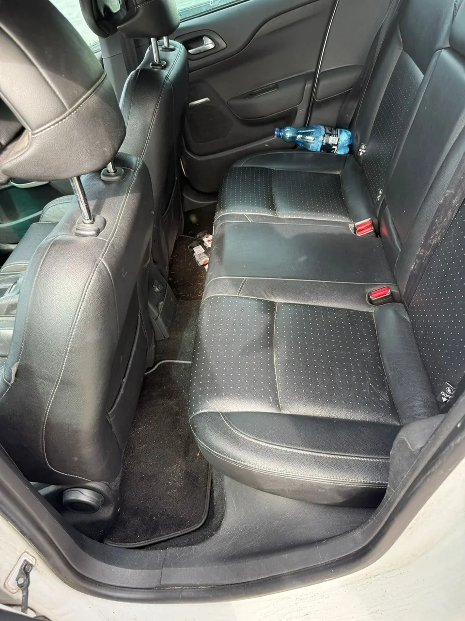 Wnętrze samochodu z widokiem na czarne skórzane siedzenia tylnego rzędu z kropkowanym wzorem. Na siedzeniu po prawej stronie leży niebieska plastikowa butelka. Widoczne są czarne podłokietniki, czerwone klamry pasów bezpieczeństwa oraz fragment drzwi z uchwytem i okienkiem. Na podłodze znajduje się czarny dywanik i rozrzucone drobne przedmioty. Siedzenia mają zagłówki, a obicie pokazuje ślady użytkowania.