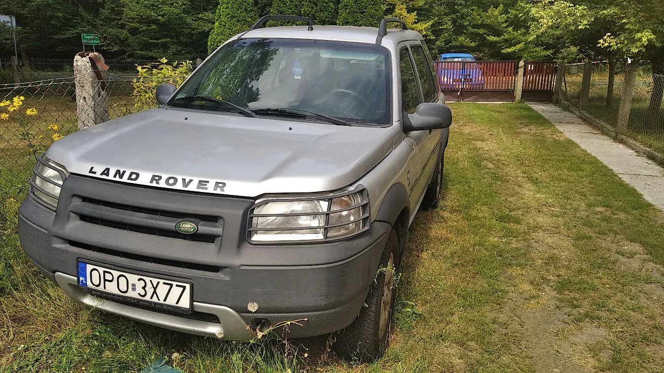 Srebrny samochód marki Land Rover zaparkowany na trawiastym podjeździe. Pojazd jest widoczny z przodu, pod kątem. Na masce widnieje napis „LAND ROVER”, a na przednim zderzaku znajduje się polska tablica rejestracyjna o numerze „OPO 3X77”. W tle widać zielone drzewa, krzewy oraz drewniany płot i taras.