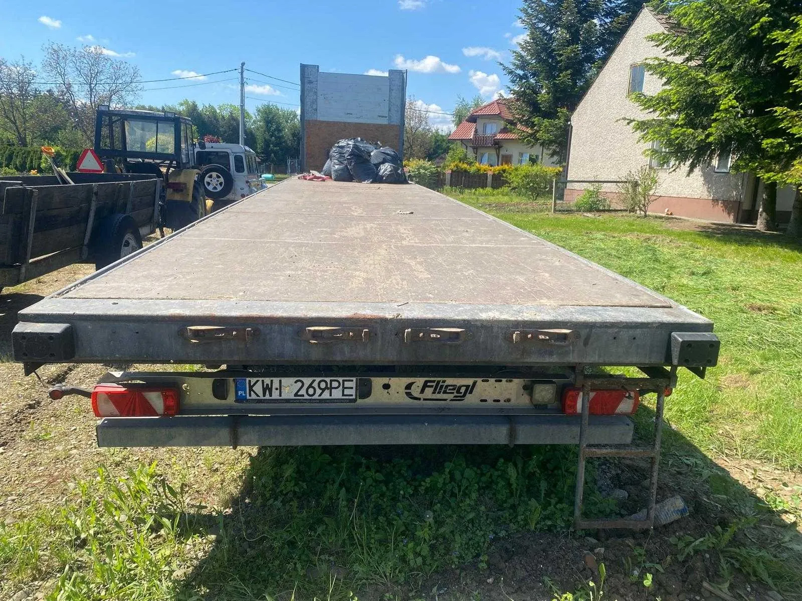 Widok z tyłu na metalową przyczepę platformową zaparkowaną na trawie. Na tylnej belce przyczepy znajduje się tablica rejestracyjna o numerze KW 269PE oraz napis 'Fliegl'. Na platformie przyczepy leżą czarne worki na śmieci. W tle widoczny jest ciągnik rolniczy, domy jednorodzinne oraz drzewa pod niebieskim niebem z lekkimi chmurami.