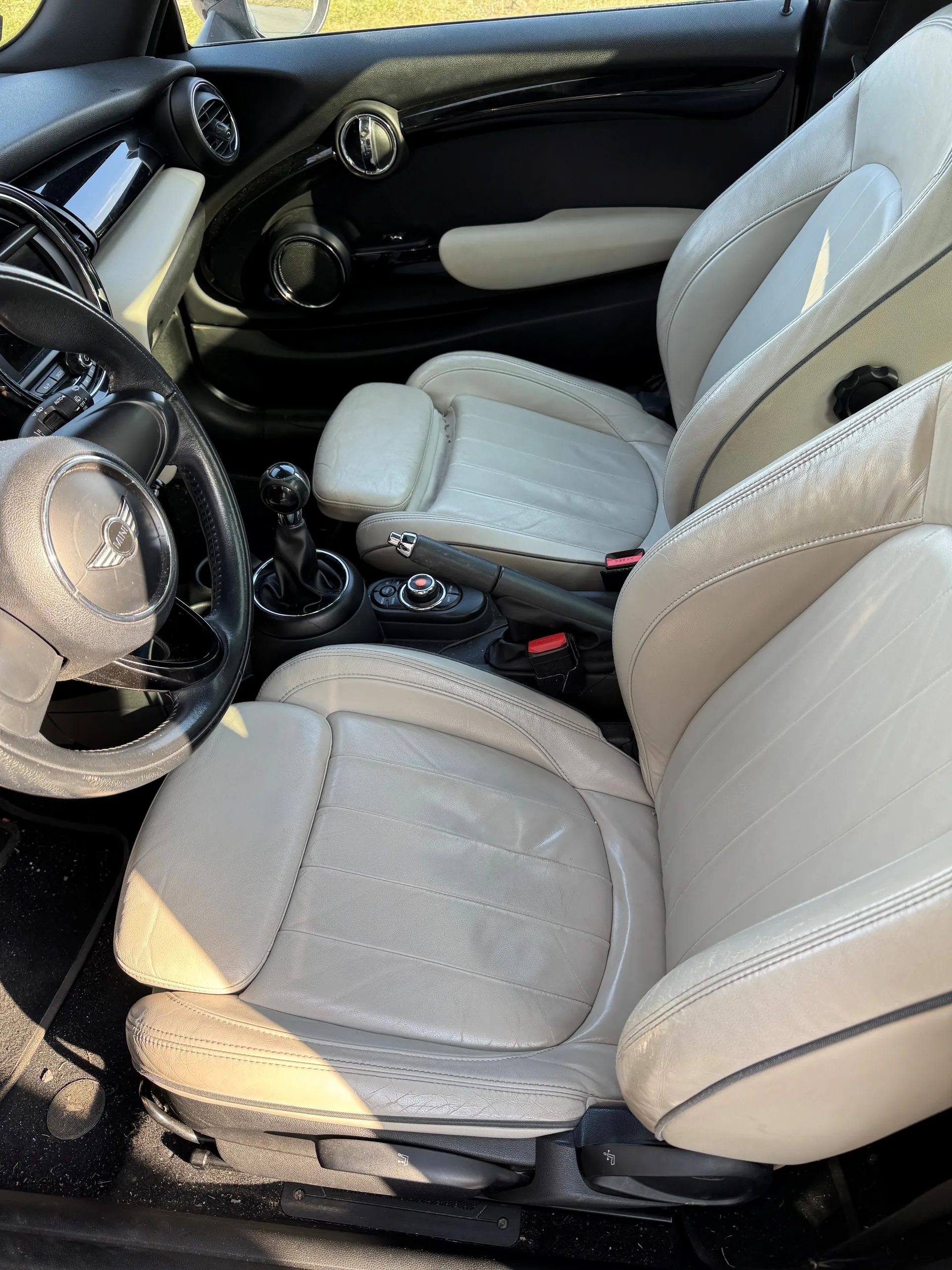 Wnętrze samochodu z widocznymi przódowymi siedzeniami w kolorze jasnoszarym, kierownicą z logo, dźwignią zmiany biegów, hamulcem ręcznym, czarną deską rozdzielczą z wentylacjami i głośnikami oraz panelami drzwiowymi z uchwytem i głośnikiem. Wnętrze oświetlone jest światłem słonecznym.