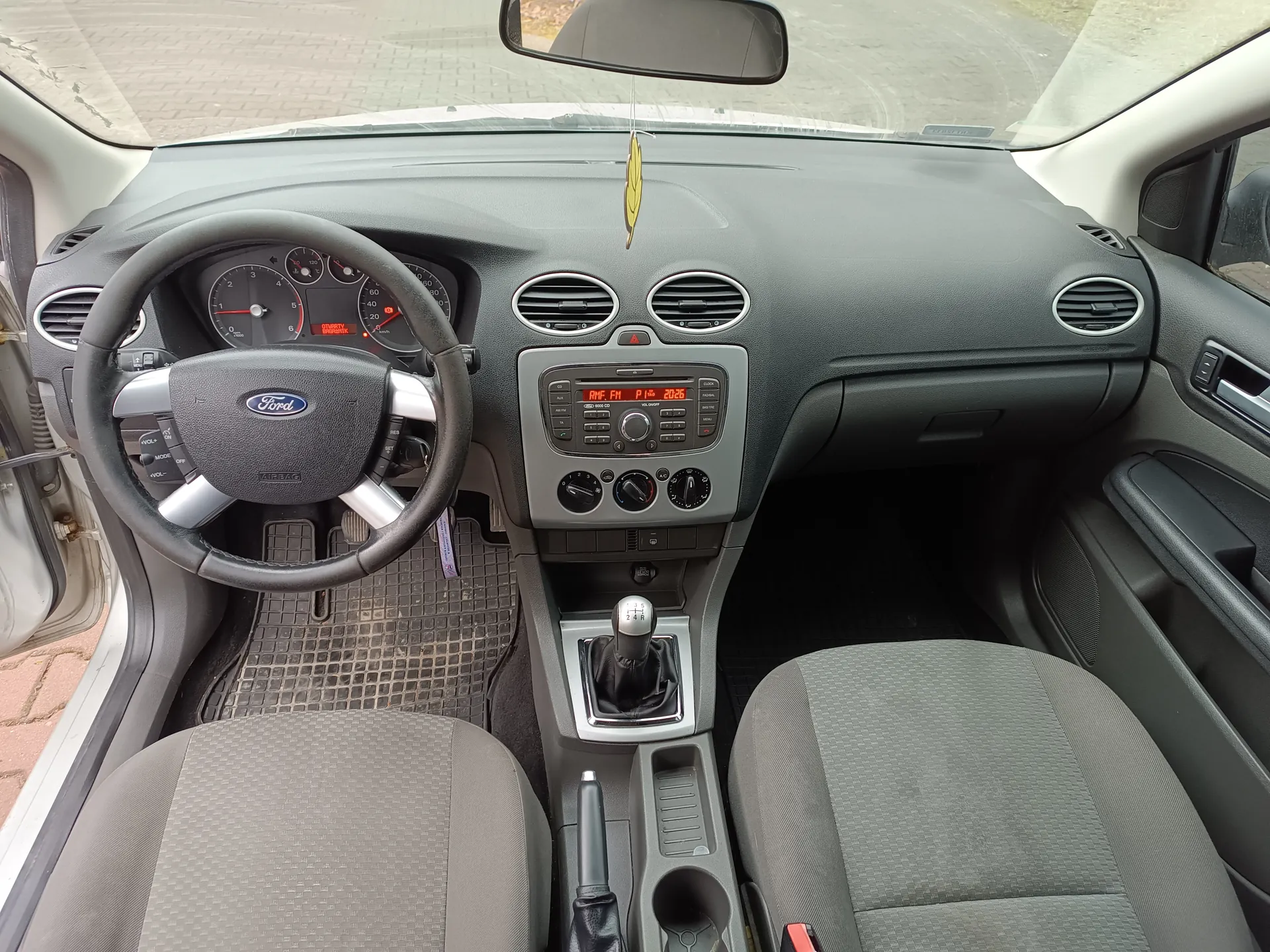 Wnętrze samochodu marki Ford z widoczną kierownicą z logo Ford, tablicą rozdzielczą z licznikami, centralnym panelem z odtwarzaczem radiowym wyświetlającym "FM 1" i "20:26", przyciskami klimatyzacji oraz dźwignią zmiany biegów. Na desce rozdzielczej wiszący żółty aromatyzator. Widoczne są dwa przednie fotele, otwarte drzwi po stronie kierowcy oraz fragment zewnętrzny przez przednią szybę z brukowaną powierzchnią.