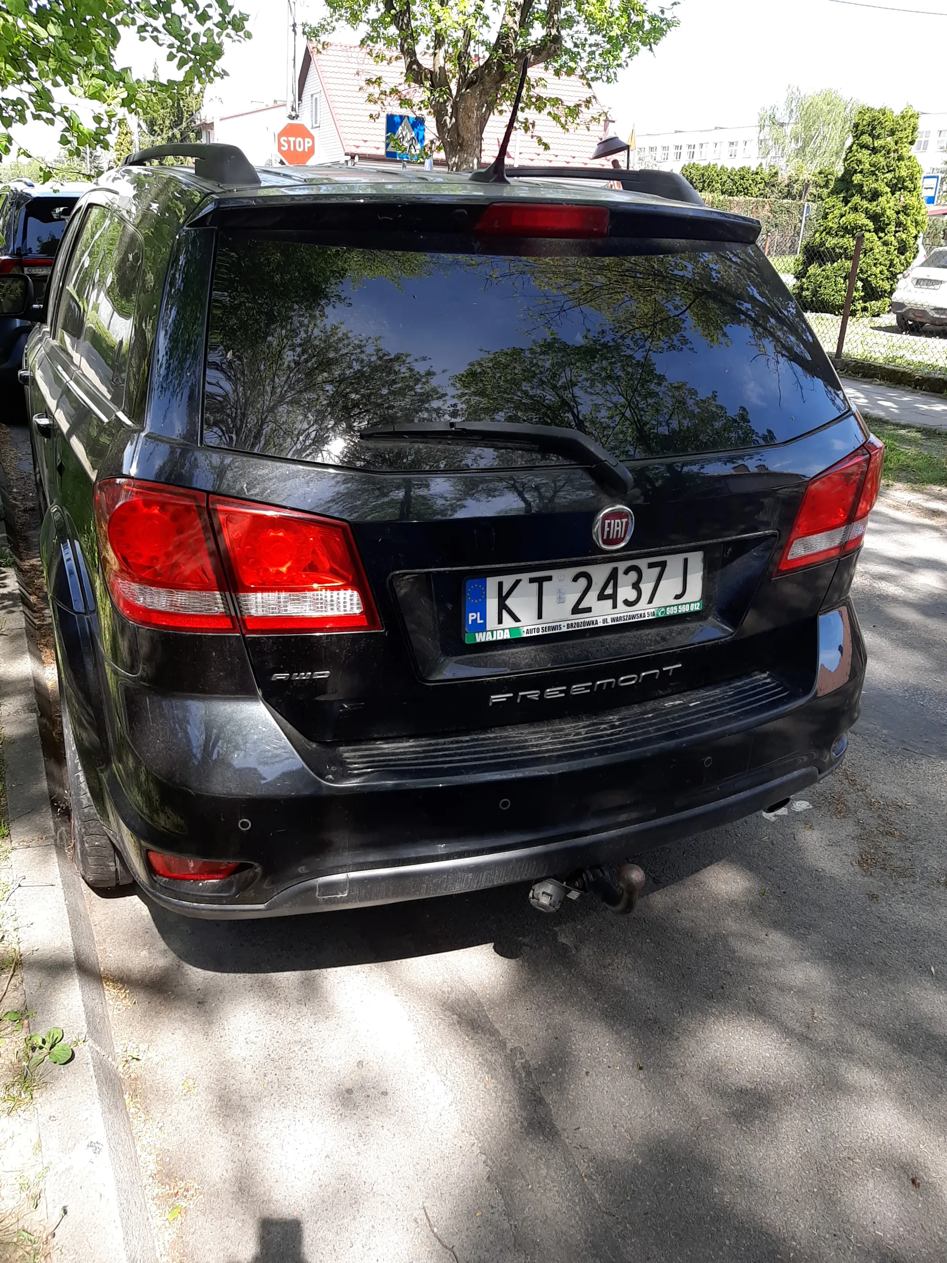 Czarny samochód Fiat Freemont zaparkowany pionowo na drodze. Tablica rejestracyjna KT 2437J, czerwone światła tylne, logo FIAT oraz napis FREEMONT na bagażniku. W tle drzewa, budynki, znak STOP oraz asfaltowa droga.