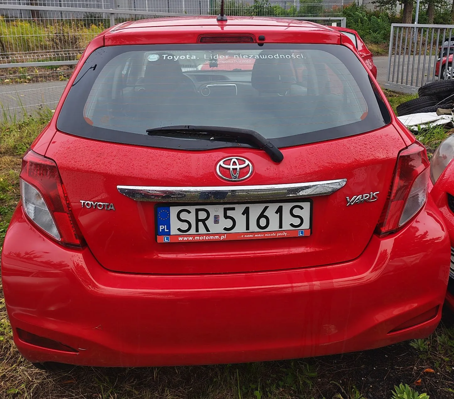 Tylny widok czerwonego samochodu marki Toyota. Na klapie bagażnika znajduje się logo Toyoty, napis 'TOYOTA' po lewej stronie oraz oznaczenie modelu 'VVT-i' po prawej. Samochód ma polską tablicę rejestracyjną o numerze SR 5161S z logo PL. Poniżej tablicy znajduje się ramka z adresem strony internetowej www.motomm.pl. Na górnej części tylnej szyby naklejona jest biała napisana informacja: 'Toyota. Lider niezawodności'.