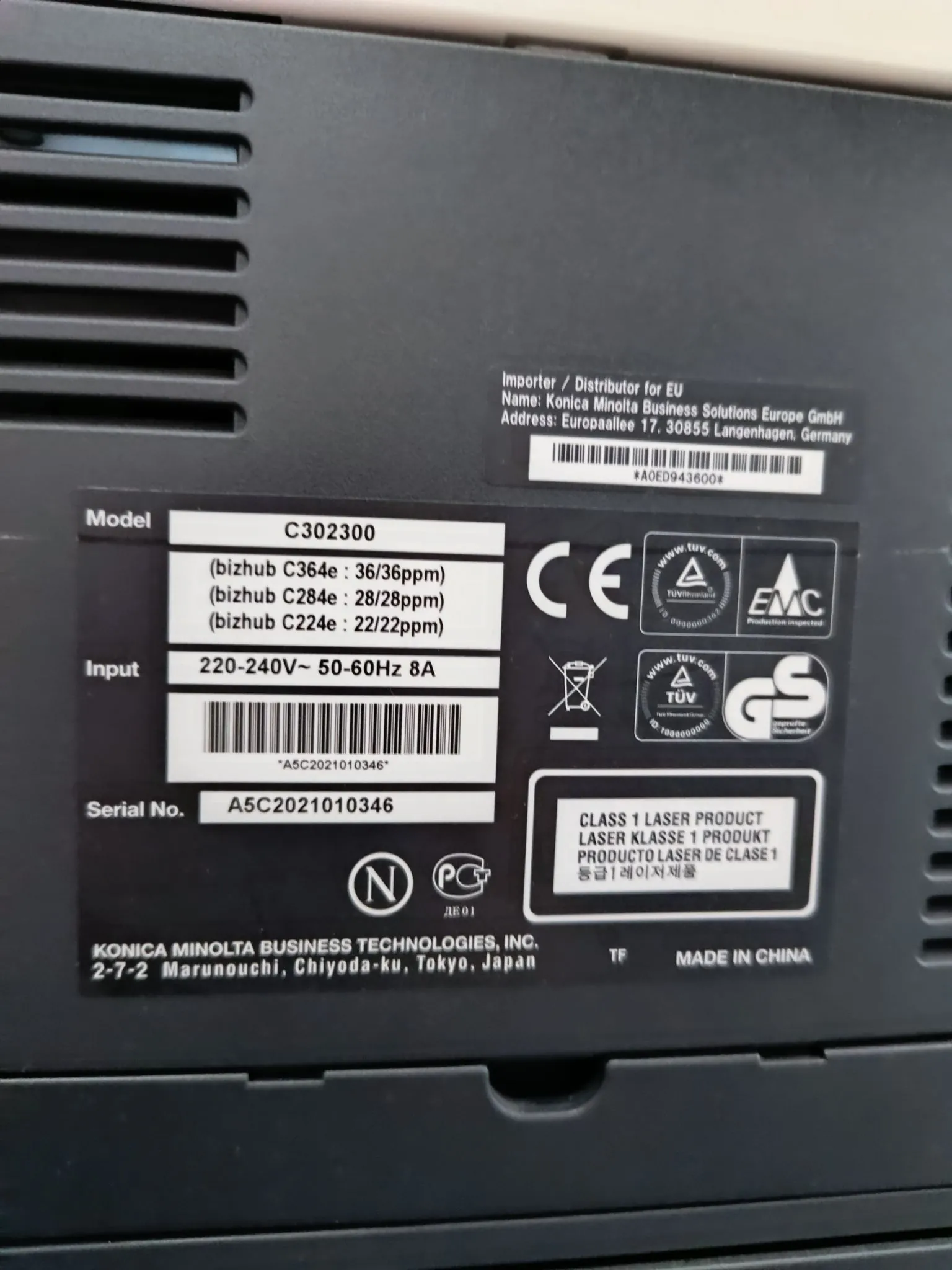 Zdjęcie tabliczki znamionowej urządzenia elektronicznej marki Konica Minolta. Na tabliczce widnieją następujące informacje: Model C302300 (bizhub C364e: 36/36ppm, bizhub C284e: 28/28ppm, bizhub C224e: 22/22ppm), Input 220-240V~ 50-60Hz 8A, Serial No. A5C2021010346. Widoczne są kody kreskowe, certyfikaty CE, EMC, GS oraz TUV. Na dole znajduje się napis: KONICA MINOLTA BUSINESS TECHNOLOGIES, INC. 2-7-2 Marunouchi, Chiyoda-ku, Tokyo, Japan oraz MADE IN CHINA. W górnej części widnieją dane importera: Konica Minolta Business Solutions Europe GmbH, Europapallee 17, 30855 Langenhagen, Germany.