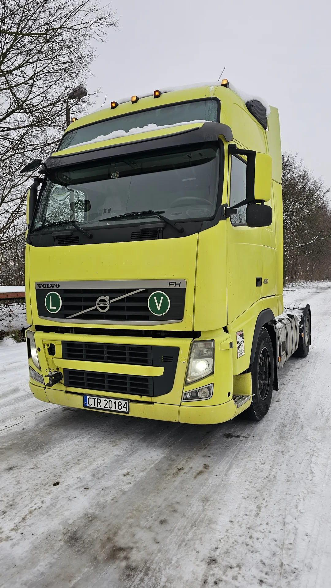 Jasnożółty ciężarówka Volvo FH z tablicą rejestracyjną CTR 20184 zaparkowana na śnieżnej, brudnej drodze. Widoczne logo Volvo, zielone okręgi z literami V i L na masce, ośnieżone szyby, włączony reflektor. W tle nagie drzewa i szare niebo.