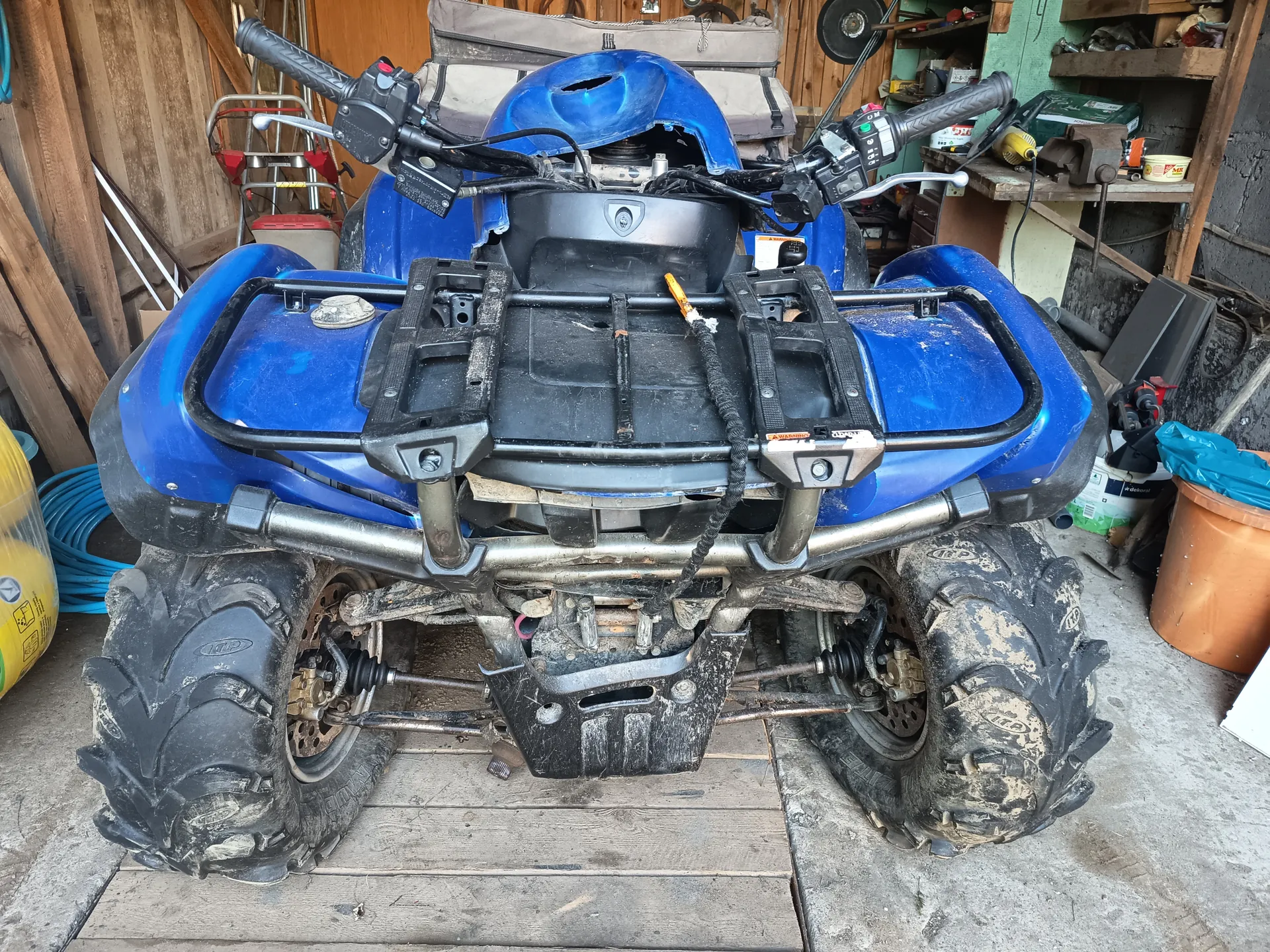 Niebieski quad z czarnym bagażnikiem i brudnymi oponami, stojący w garażu. W tle widoczne są drewniane ściany, narzędzia na półkach, pomarańczowy wiadro, niebieskie wężo i żółty pojemnik. Quad ma czarne uchwyty, elementy metalowe i widoczne ślady użytkowania. Na oponach widoczny napis 'ITP'.
