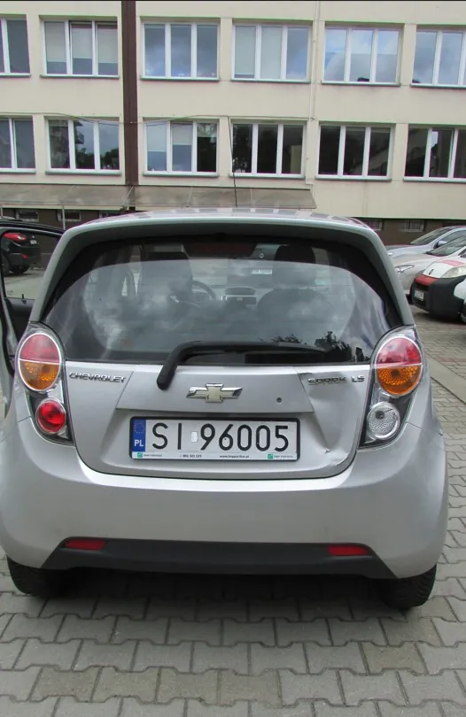 Tylny widok srebrnego samochodu marki Chevrolet Spark. Na klapie bagażnika widoczne jest logo marki, napis 'CHEVROLET' po lewej stronie oraz 'SPARK LS' po prawej. Samochód posiada polską tablicę rejestracyjną o numerze SI 96005. Auto zaparkowane jest na kostce brukowej przed jasnym budynkiem z wieloma oknami.