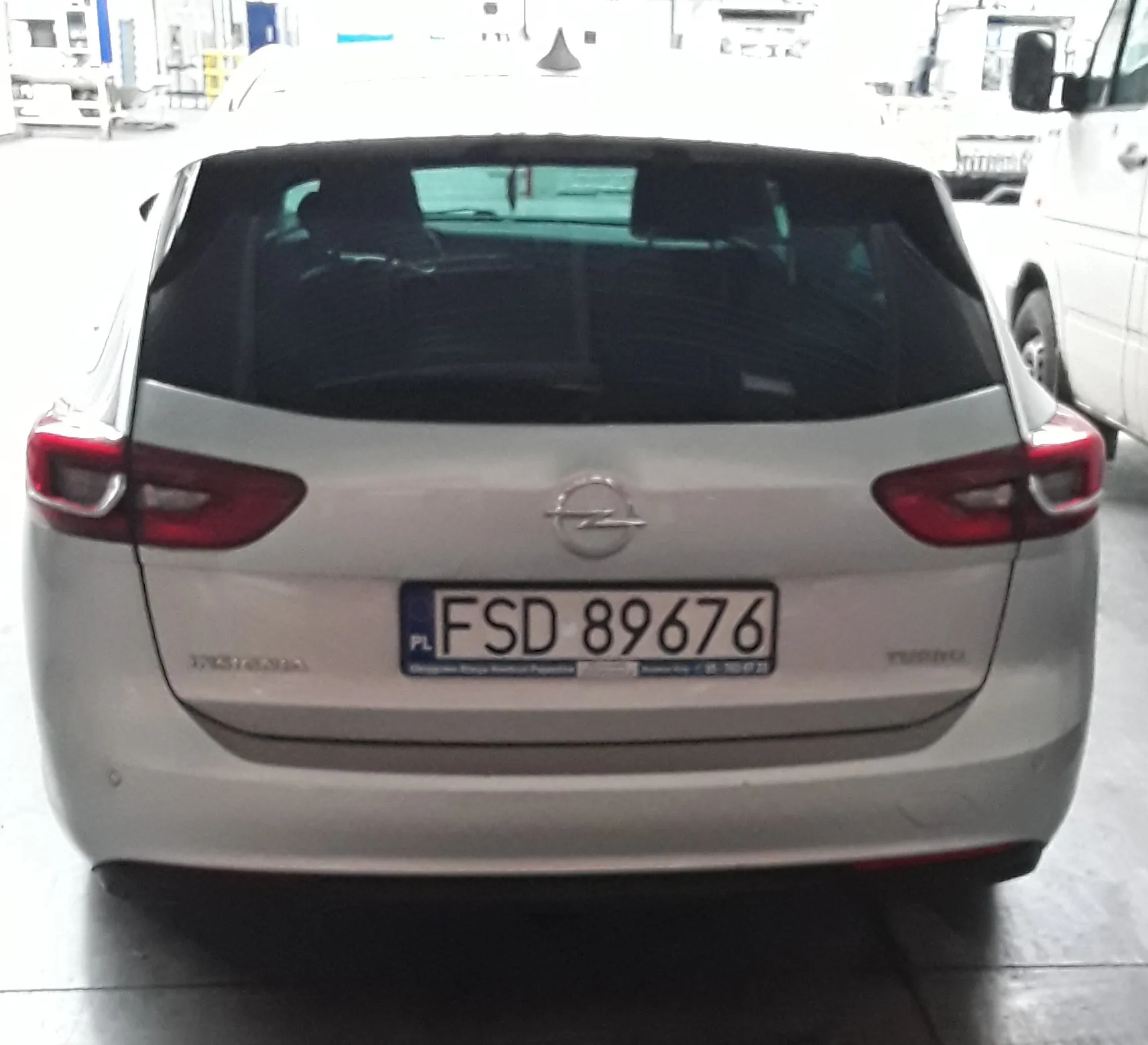 Tylny widok srebrnego samochodu marki Opel. Na klapie bagażnika znajduje się logo producenta oraz polska tablica rejestracyjna o numerze FSD 89676. Po lewej stronie widnieje napis INSIGNIA, a po prawej napis TURBO.