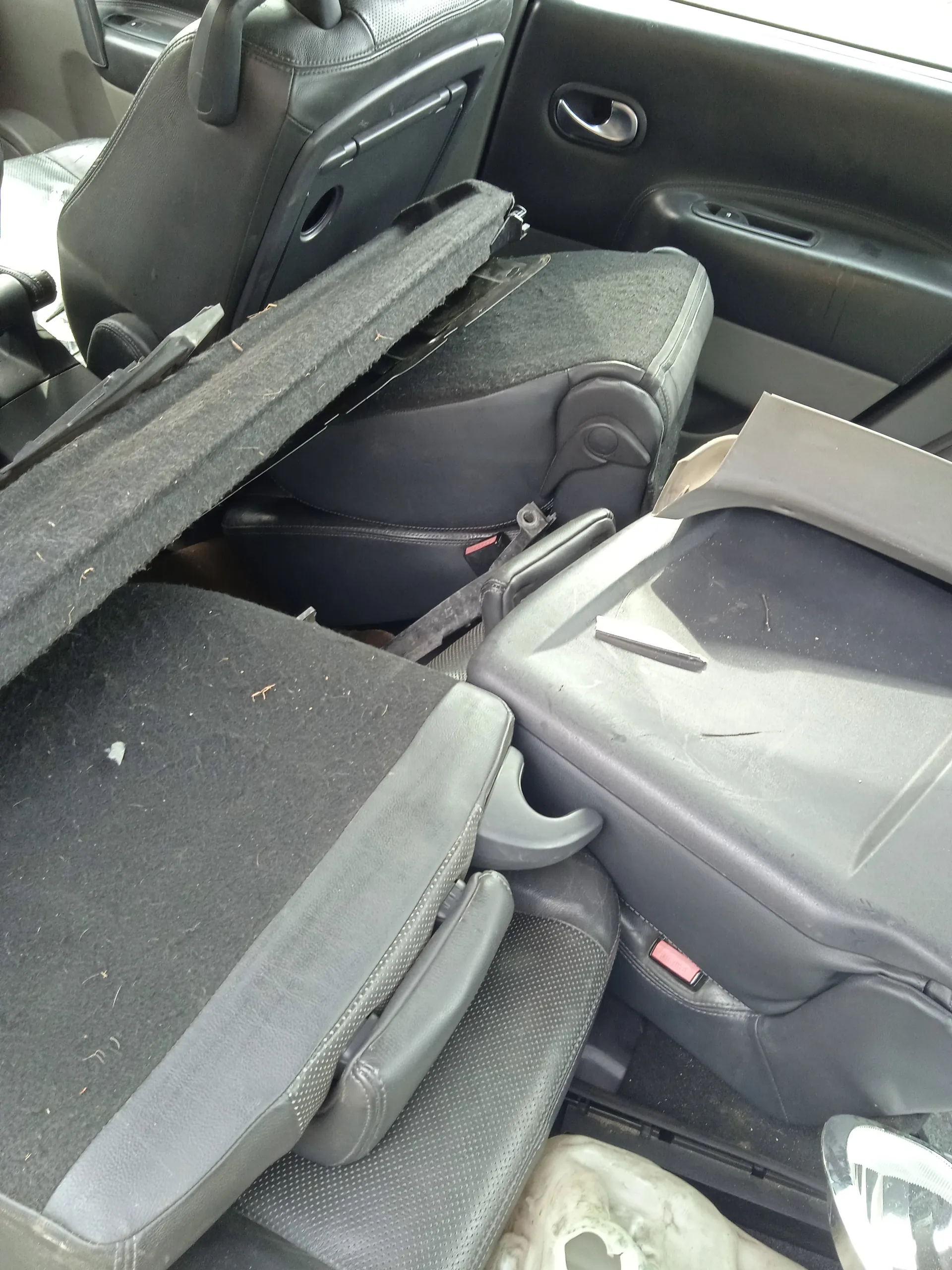 Zdjęcie przedstawia wnętrze samochodu z czarnymi skórzonymi fotelami. Widoczne są przednie fotele, część z nich wydaje się być rozmontowana lub przesunięta. Na siedzeniach leży czarna poduszka lub fragment wykładziny z widocznymi śladami brudu i drobnymi szczątkami. W tle widoczna jest czarna deska rozdzielcza z uchwytem drzwiowym oraz fragmenty pasów bezpieczeństwa. Na środkowej konsoli znajduje się mały, ciemny przedmiot przypominający długopis. Wnętrze samochodu wydaje się nieporządne, z widocznymi śladami zużycia.