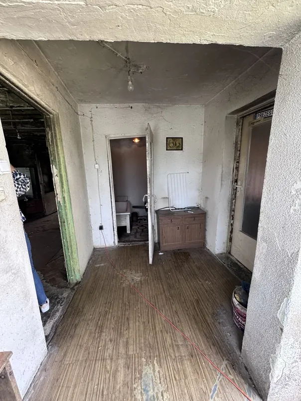 Wnętrze zniszczonego pomieszczenia przypominającego korytarz. Podłoga wyłożona jest brązowym linoleum w paski, przez które przebiega czerwony kabel elektryczny. Po lewej stronie widać fragment osoby w niebieskich spodniach i białych butach. W głębi znajduje się otwarte przejście do kolejnego pokoju, w którym widoczne jest białe krzesło. Po prawej stronie stoi brązowa szafka z szufladami, nad nią znajduje się biały grzejnik i mały obrazek w ramce na ścianie. Po prawej stronie widoczne są drzwi z napisem 'PREZEST' na górnej ramie. Ściany są odrapane, z widocznymi ubytkami tynku.