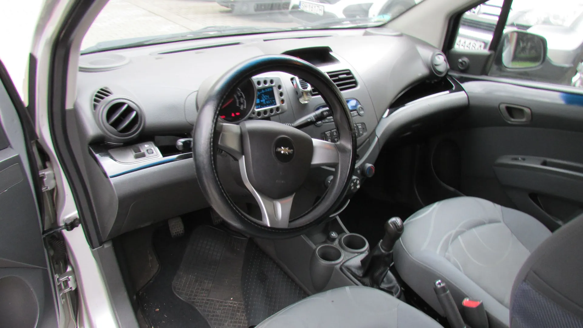 Wnętrze samochodu marki Chevrolet widziane z perspektywy otwartych drzwi kierowcy. Widoczna jest czarna kierownica z logo producenta, deska rozdzielcza z zegarami, nawiewy powietrza, lewarek zmiany biegów oraz szare fotele materiałowe.
