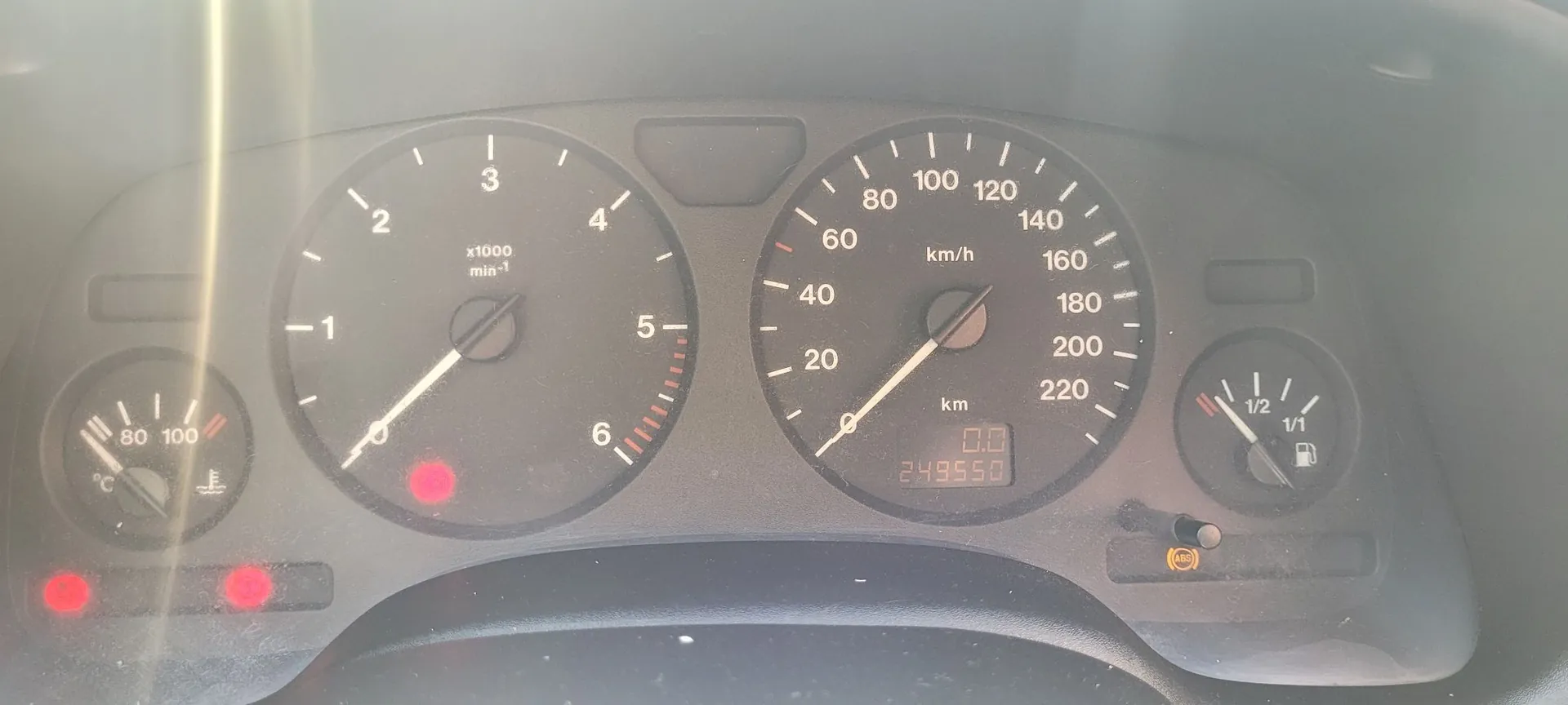 Panel instrumentów w samochodzie. Po lewej stronie znajduje się wskaźnik temperatury cieczy chłodzącej z oznaczeniami 80 i 100 stopni Celsjusza. W centrum po lewej jest obrotomierz z oznaczeniami od 1 do 6 x1000 min-1. Po prawej stronie znajduje się prędkościomierz z podziałką od 0 do 220 km/h oraz cyfrowym licznikiem przebiegu wskazującym wartość 249550 km. Po prawej stronie znajduje się wskaźnik poziomu paliwa z oznaczeniami 1/2 i 1/1. Na panelu widoczne są zapalone czerwone kontrolki ostrzegawcze oraz żółta kontrolka ABS.