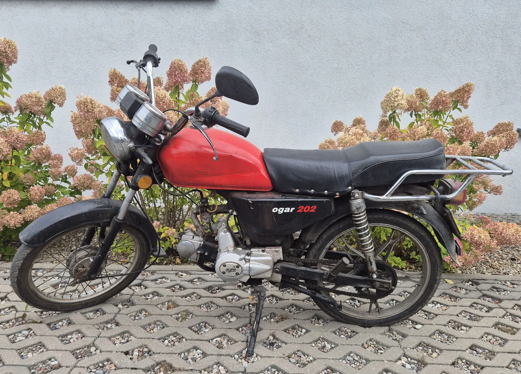 Czerwony motocykl marki Ogar 202 zaparkowany na kostce brukowej na tle jasnej ściany i krzewów hortensji. Pojazd ma czarne siedzenie, srebrne felgi szprychowe i widoczny napis 'ogar 202' na bocznej osłonie silnika.