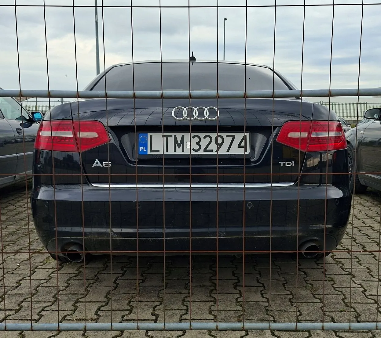 Tylny widok czarnego samochodu marki Audi A6 TDI z polską tablicą rejestracyjną LTM 32974. Samochód znajduje się za metalowym ogrodzeniem z drutu. W tle widać zachmurzone niebo oraz fragmenty innych pojazdów.