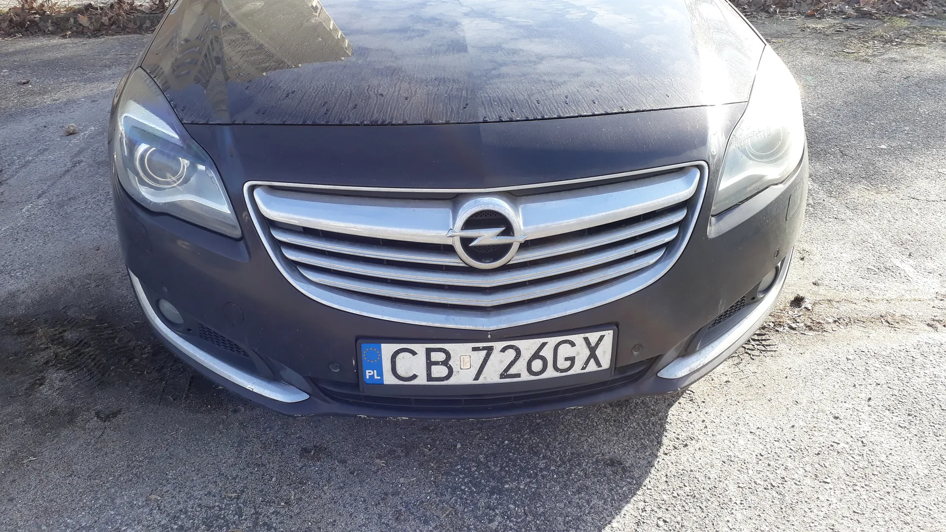 Przód ciemnoszarego samochodu marki Opel z srebrną atrapą chłodnicy i logo w kształcie litery "Z" w okręgu. Widoczna tablica rejestracyjna z białym tłem, czarnymi literami i cyframi "CB 726GX" oraz niebieskim pasem z napisem "PL" po lewej stronie. Samochód zaparkowany na asfaltowej nawierzchni, w tle suche liście i fragment chodnika.