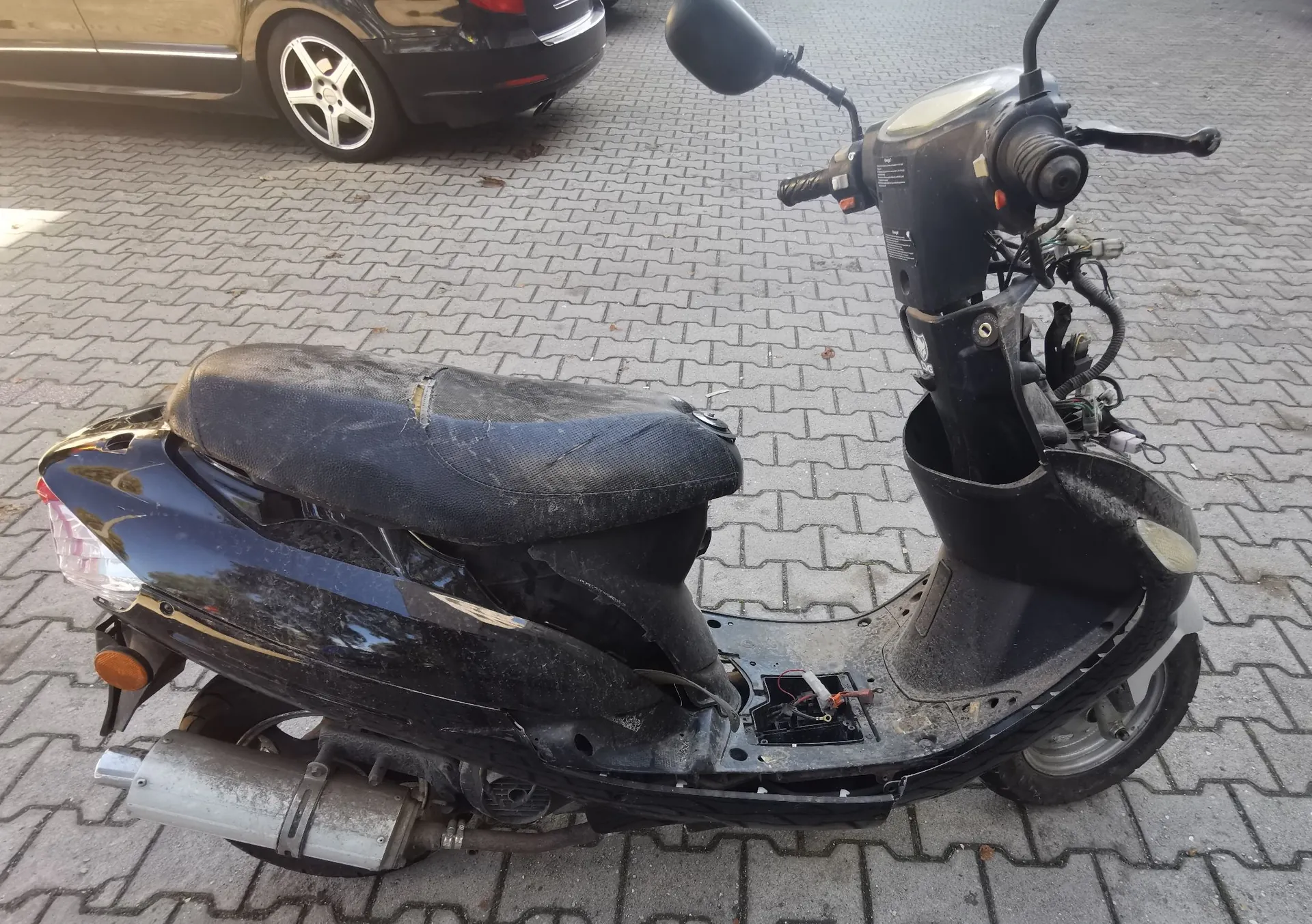 Czarny skuter zaparkowany na kostce brukowej. Pojazd jest w złym stanie technicznym: siedzenie jest przetarte i popękane, brakuje wielu elementów plastikowych osłon w przedniej części, a przewody elektryczne są odsłonięte. W tle widoczny jest fragment czarnego samochodu osobowego.