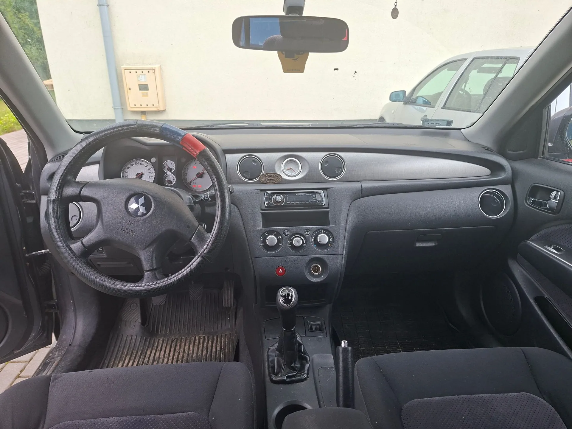 Wnętrze samochodu marki Mitsubishi widziane z perspektywy przednich siedzeń. Widoczna jest czarna kierownica z logo producenta, deska rozdzielcza z analogowymi wskaźnikami, pokrętłami klimatyzacji oraz radiem. W centralnej części znajduje się dźwignia zmiany biegów. Przez przednią szybę widoczny jest fragment białego budynku oraz zaparkowany biały samochód.