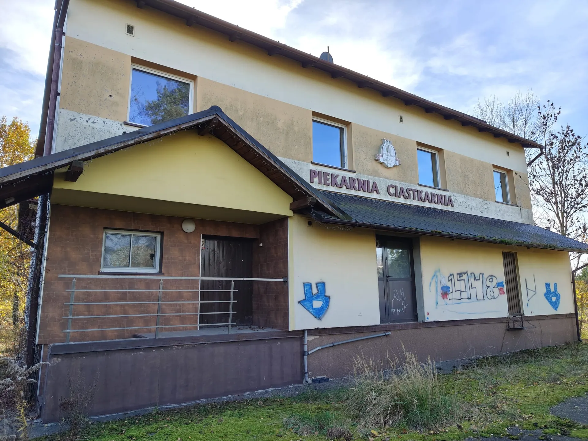 Dwukondygnacyjny budynek o żółtej elewacji z ciemnobrązowym fundamentem i dachem. Na fasadzie znajduje się napis 'PIEKARNIA CIASTKARNIA'. Parter budynku posiada ciemnobrązowe drzwi wejściowe, okno oraz fragmenty graffiti w kolorach niebieskim i czerwonym. Na piętrze widoczne są cztery prostokątne okna oraz mały emblemat nad napisem. Przed budynkiem znajduje się trawnik z kępami suchej trawy.