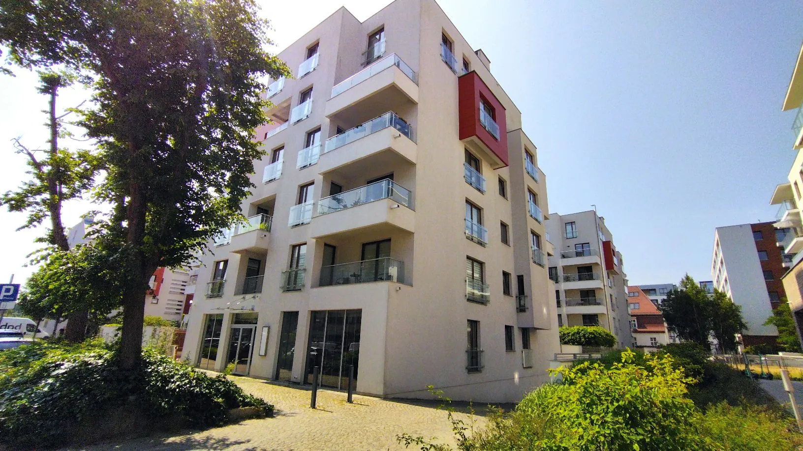 Nowoczesny, wielopiętrowy budynek mieszkalny o jasnej elewacji z licznymi balkonami z szklanymi barierkami. Część fasady posiada czerwony akcent architektoniczny w formie wystającego modułu. Na parterze znajdują się przeszklone witryny lokali użytkowych. Po lewej stronie zdjęcia rośnie duże, zielone drzewo, a w prawym dolnym rogu widoczne są niskie krzewy. W tle widać inne budynki mieszkalne pod bezchmurnym, jasnym niebem.