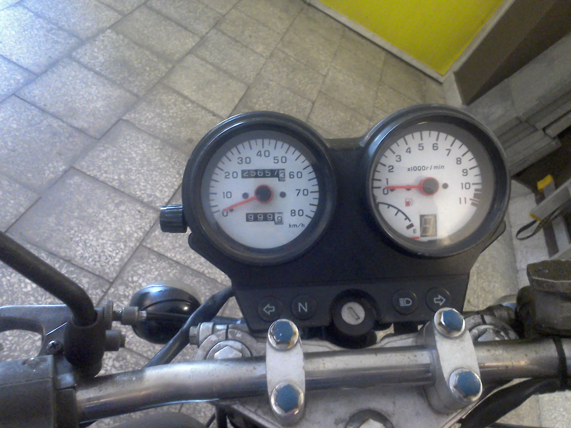 Widok z perspektywy kierowcy na deskę rozdzielczą motocykla. Widoczne są dwa okrągłe wskaźniki: po lewej prędkościomierz z podziałką do 100 km/h i licznikiem przebiegu, po prawej obrotomierz z podziałką do 12 x1000 r/min. Poniżej zegarów znajduje się panel z kontrolkami, w tym oznaczeniem biegu neutralnego 'N'. W dolnej części zdjęcia widoczna jest srebrna kierownica z uchwytami i śrubami montażowymi. Tło stanowi szara, płytkowa nawierzchnia.**