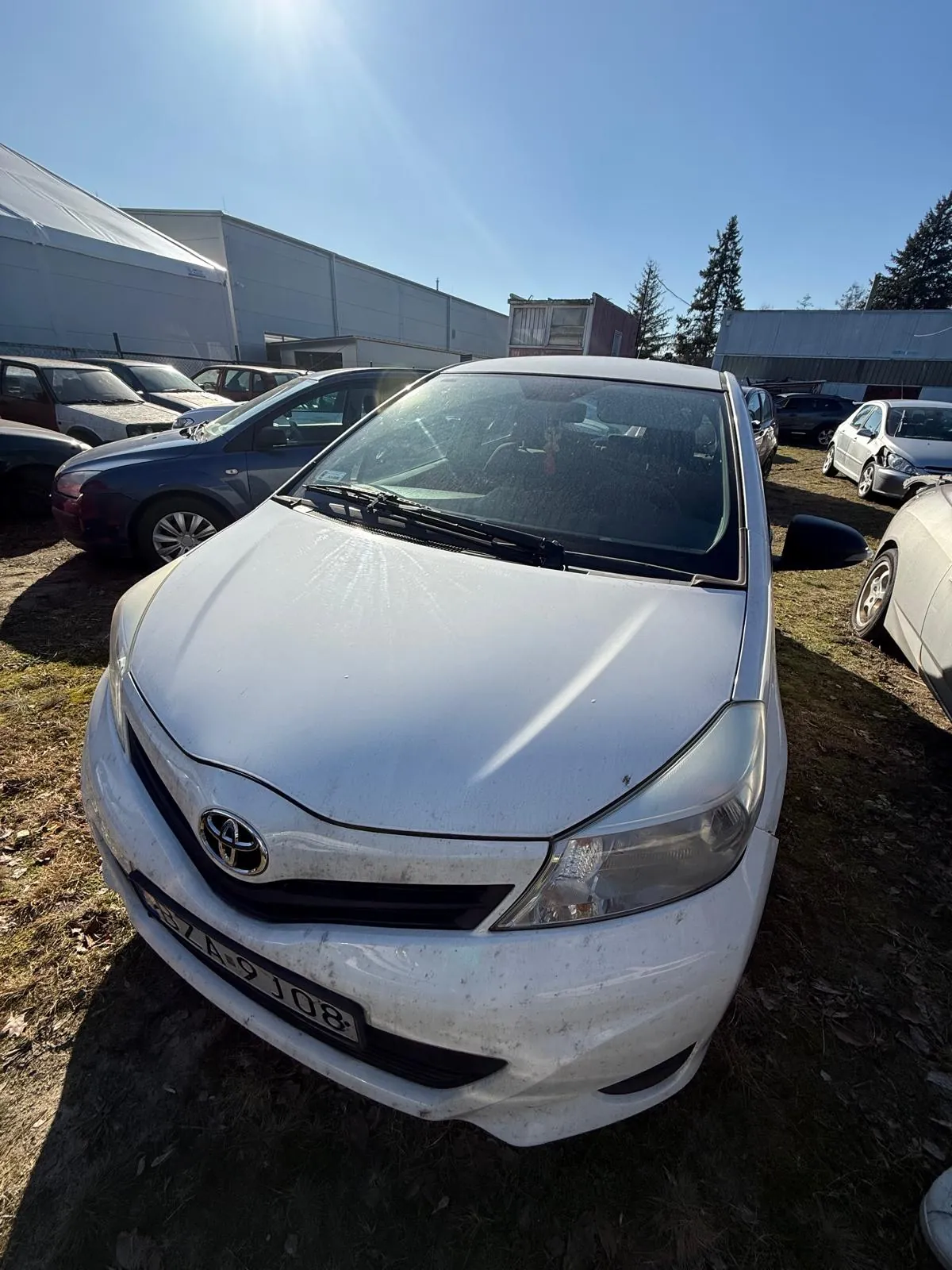 Biały samochód marki Toyota z tablicą rejestracyjną 37A 9J08 na pierwszym planie. W tle widoczne są inne zaparkowane pojazdy, budynek przemysłowy, namiot oraz drzewa. Niebo jest bezchmurne, słońce świeci jasno. Podłoże pokryte jest suchą trawą i liśćmi.