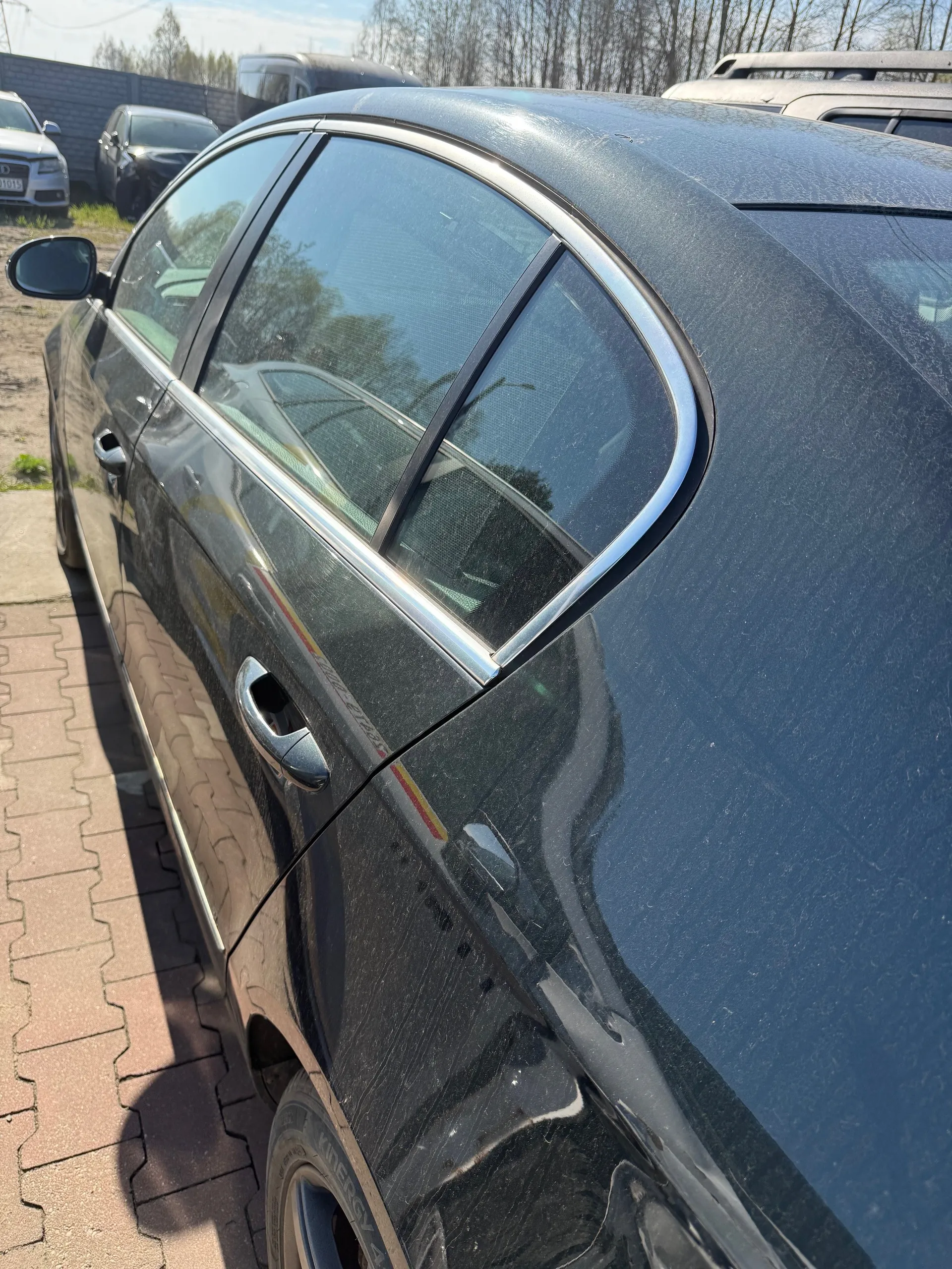 Czarny samochód osobowy typu sedan, widok z boku od tyłu. Pojazd ma połyskującą karoserię z chromowaną listwą wzdłuż okien i przyciemnionymi szybami. W tle widoczne są inne zaparkowane samochody, brukowana powierzchnia, fragment trawnika, drzewa oraz betonowy płot. Na szybach odbijają się otoczenie i inne pojazdy. Niebo jest jasne, słoneczna pogoda.