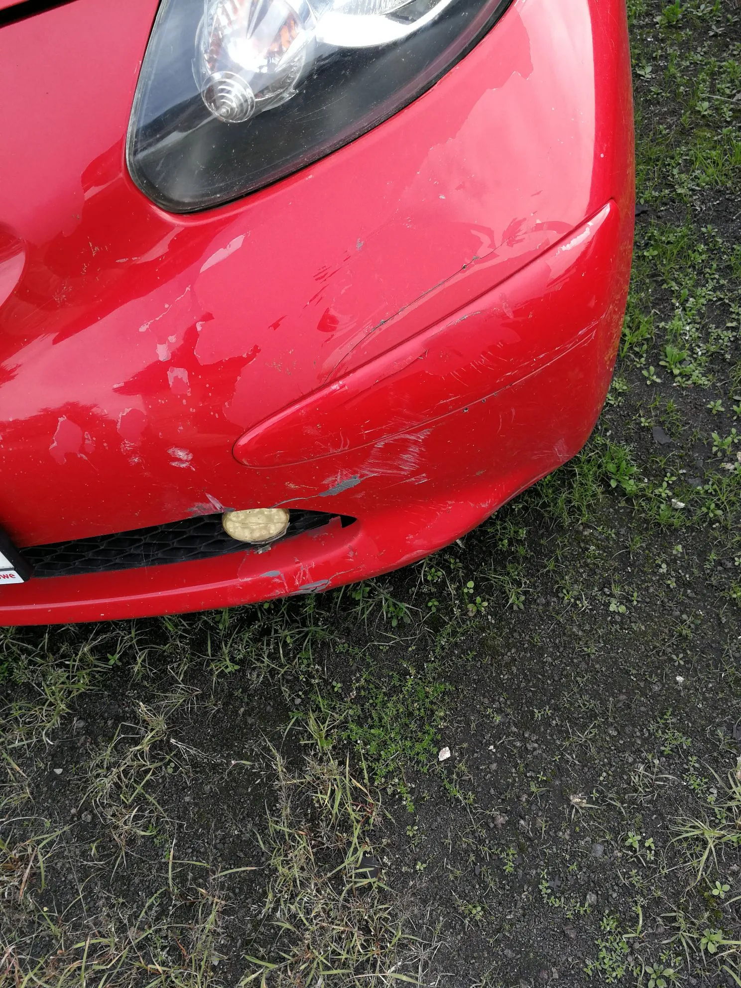 Zbliżenie na przedni, czerwony zderzak samochodu z widocznymi uszkodzeniami w postaci licznych zarysowań, otarć oraz odprysków lakieru. W górnej części widoczny jest fragment reflektora, a poniżej mała, okrągła lampka przeciwmgielna. Samochód stoi na podłożu z ciemnej ziemi i rzadkiej trawy.