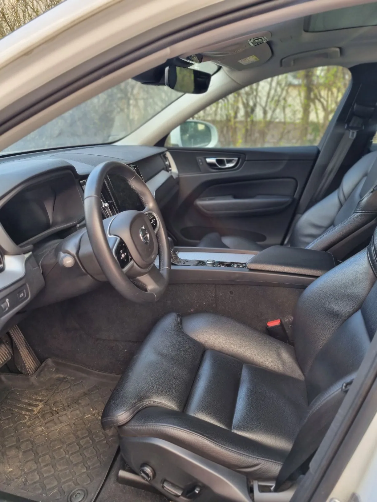 Wnętrze samochodu z czarnymi skórzonymi fotelami, kierownicą z logo, przekładnią biegów, panelami drzwiowymi i lusterkiem bocznym widocznym przez otwarte drzwi. Za oknem drzewa i inny pojazd. Na podłodze czarne maty.