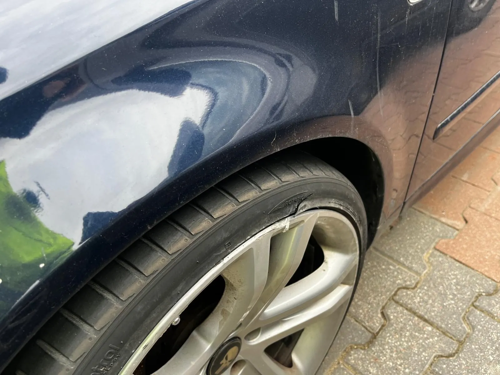 Zbliżenie na przednie koło ciemnoniebieskiego samochodu. Widoczna czarna opona z wyraźnymi rysami i częściowo czytelnym napisem 'Control', srebrna felga z logo w kształcie czaszki w środku. Nad kołem fragment błotnika z odbiciem nieba i zieleni. Tło stanowi bruk z czerwono-szarych płytek.