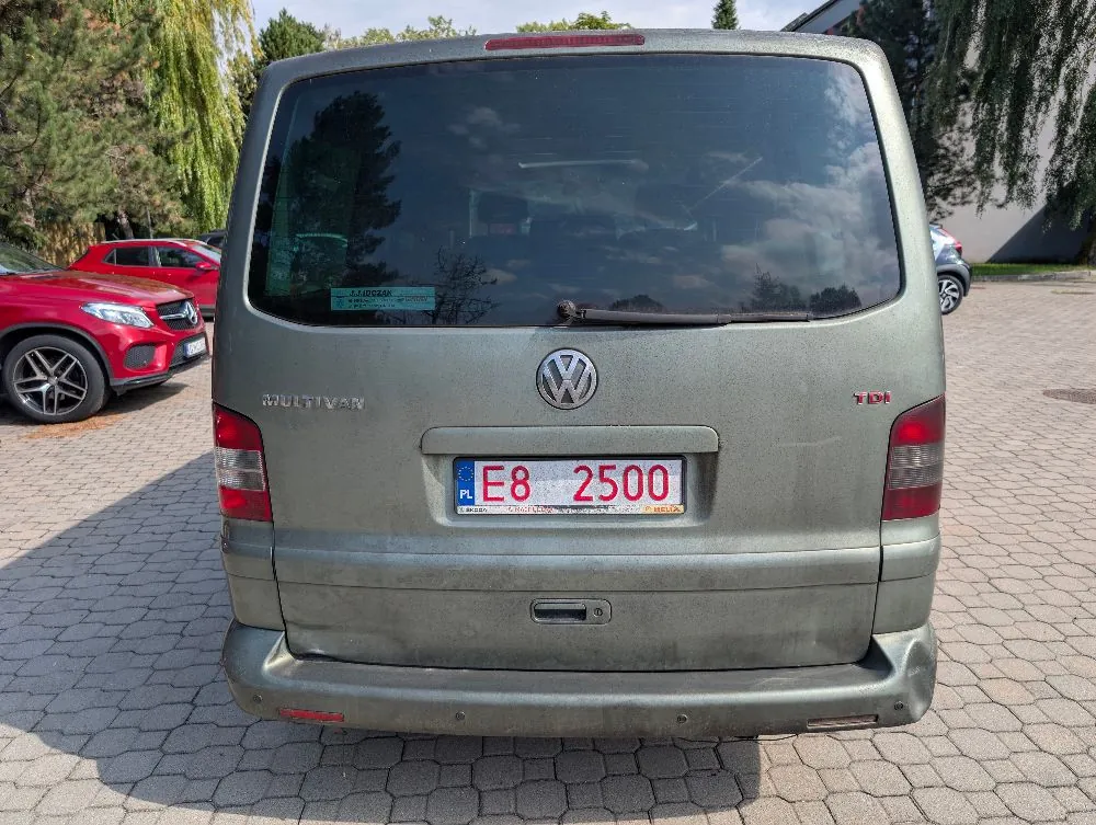 Tylny widok srebrno-szarego vana marki Volkswagen Multivan. Na klapie bagażnika widoczne jest logo Volkswagena, napis MULTIVAN po lewej stronie oraz TDI po prawej. Samochód posiada polską tablicę rejestracyjną o numerze E8 2500. W tle po lewej stronie stoi czerwony samochód osobowy, a całość znajduje się na kostce brukowej.