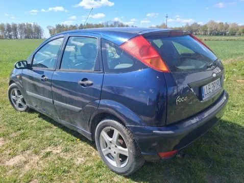 Ciemnoniebieski samochód osobowy Ford Focus pięciodrzwiowy zaparkowany na trawie. Widok z tyłu i boku. Na tablicy rejestracyjnej widoczny numer UE 3912. W tle pola, drzewa i niebo z chmurami. Na tylnym zderzaku widoczny napis focus.