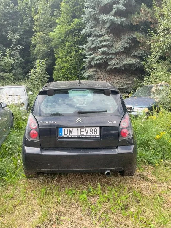 Tylny widok czarnego samochodu marki Citroën C2 zaparkowanego na trawiastym terenie. Na klapie bagażnika widoczne są napisy 'CITROËN' oraz 'C2', a także logo marki. Samochód posiada polską tablicę rejestracyjną o numerze DW 1EV88. W tle widać gęste, zielone drzewa i krzewy oraz fragmenty innych samochodów po obu stronach.