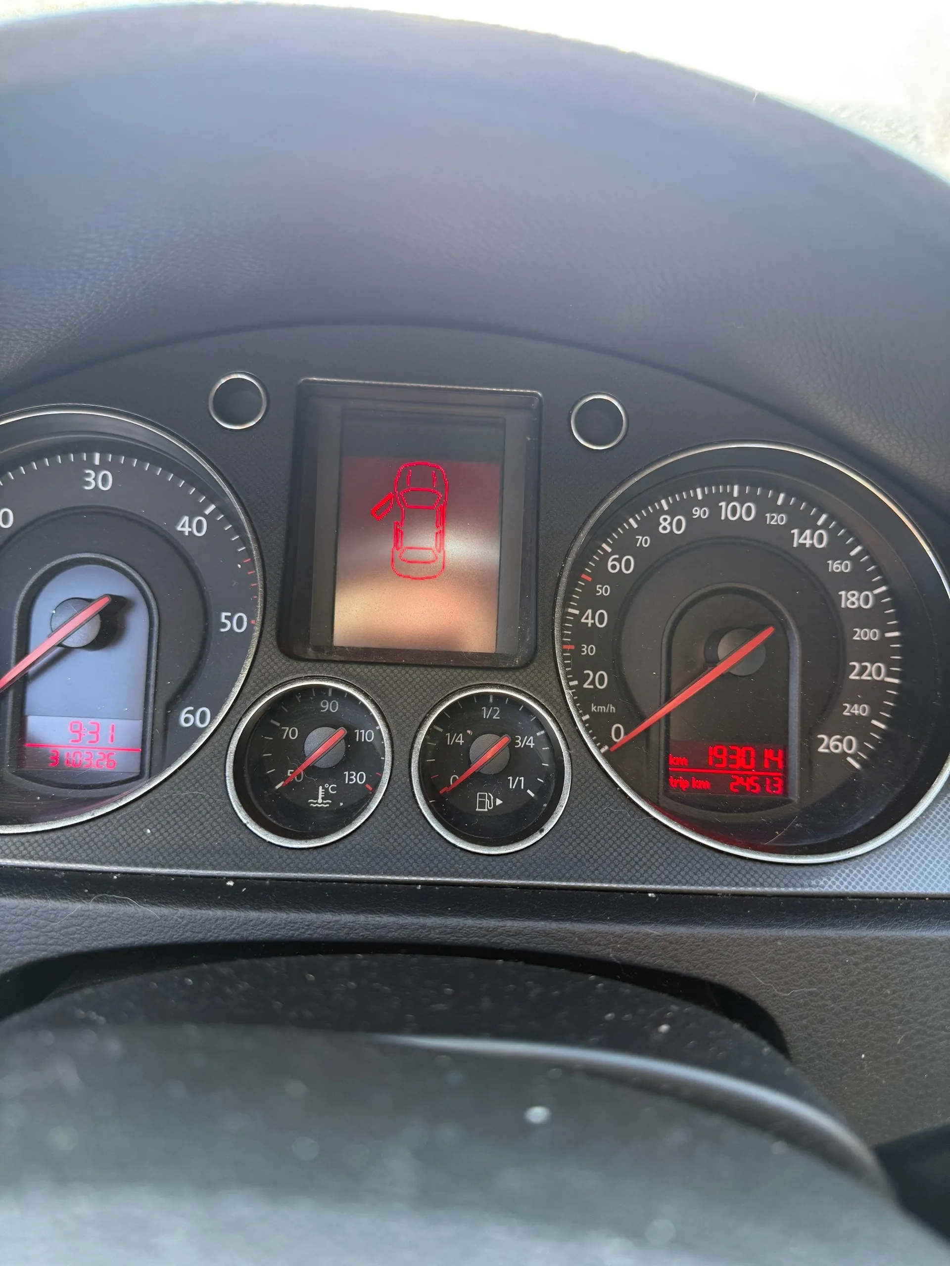 Zbliżenie na deski rozdzielcze samochodu. Po lewej stronie wskaźnik obrotomieru z czerwoną igłą, poniżej cyfrowy wyświetlacz z czerwonymi cyframi 9:31 i 310326. W centrum mały ekran z czerwonym symbolem samochodu z otwartą klapą bagażnika. Po prawej stronie wskaźnik prędkości do 260 km/h z czerwoną igłą, poniżej cyfrowy wyświetlacz z napisem "km 1930.14" i "trip km 245.13". Poniżej centralnego ekranu dwa mniejsze wskaźniki: termometr (50–130°C) i wskaźnik paliwa (1/4–1). Wszystkie cyfry i symbole są czerwone, tło deski rozdzielczej czarne. Na dole widoczny fragment kierownicy.