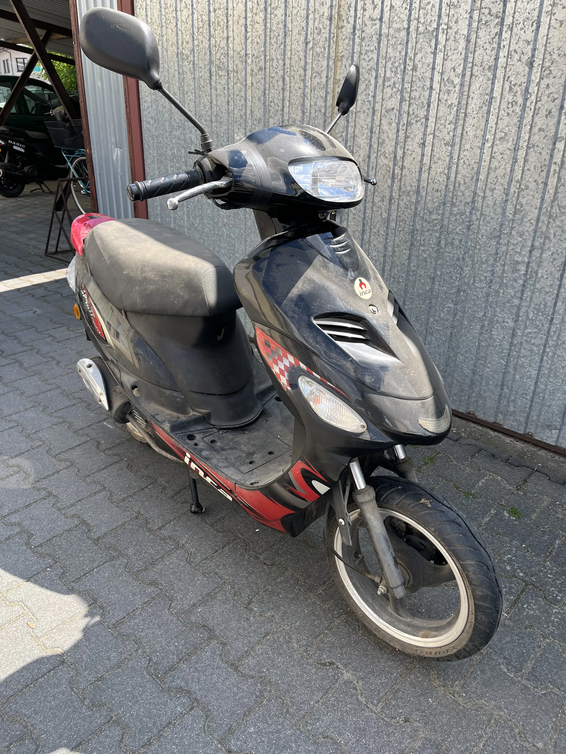Czarny skuter z czerwonymi i białymi szachownicowymi wzorami po bokach, zaparkowany na brukowanej powierzchni. Skuter posiada czarne siedzenie, reflektory, lusterka boczne oraz widoczny znak na przedniej części. W tle metalowa brama garażowa i fragment innych pojazdów. Skuter stoi na podstawce, a opona przedniego koła jest częściowo zabrudzona.