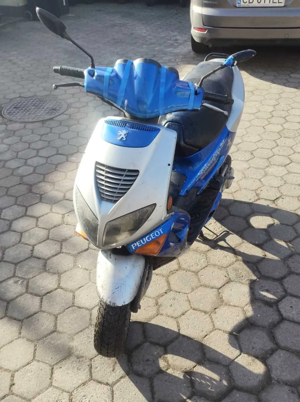 Niebiesko-biały skuter marki Peugeot z widocznym logo i napisem 'PEUGEOT' na przodzie. Na kierownicy znajduje się niebieska taśma. Skuter zaparkowany na chodniku z sześciokątnymi płytami brukowymi. W tle widoczny samochód z tablicą rejestracyjną 'CD 011EL' oraz pokrywa studzienki.