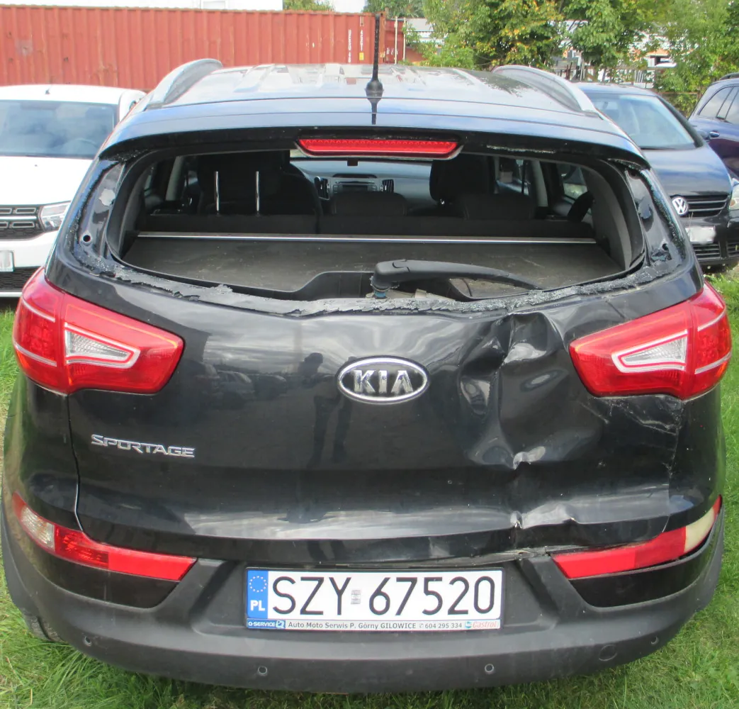 Tylny widok czarnego samochodu marki Kia Sportage z poważnymi uszkodzeniami karoserii. Tylna szyba jest całkowicie rozbita, a klapa bagażnika jest mocno wgnieciona po prawej stronie. Samochód posiada polską tablicę rejestracyjną o numerze SZY 67520. Pojazd stoi na trawie, w tle widoczne są inne samochody oraz czerwony kontener.