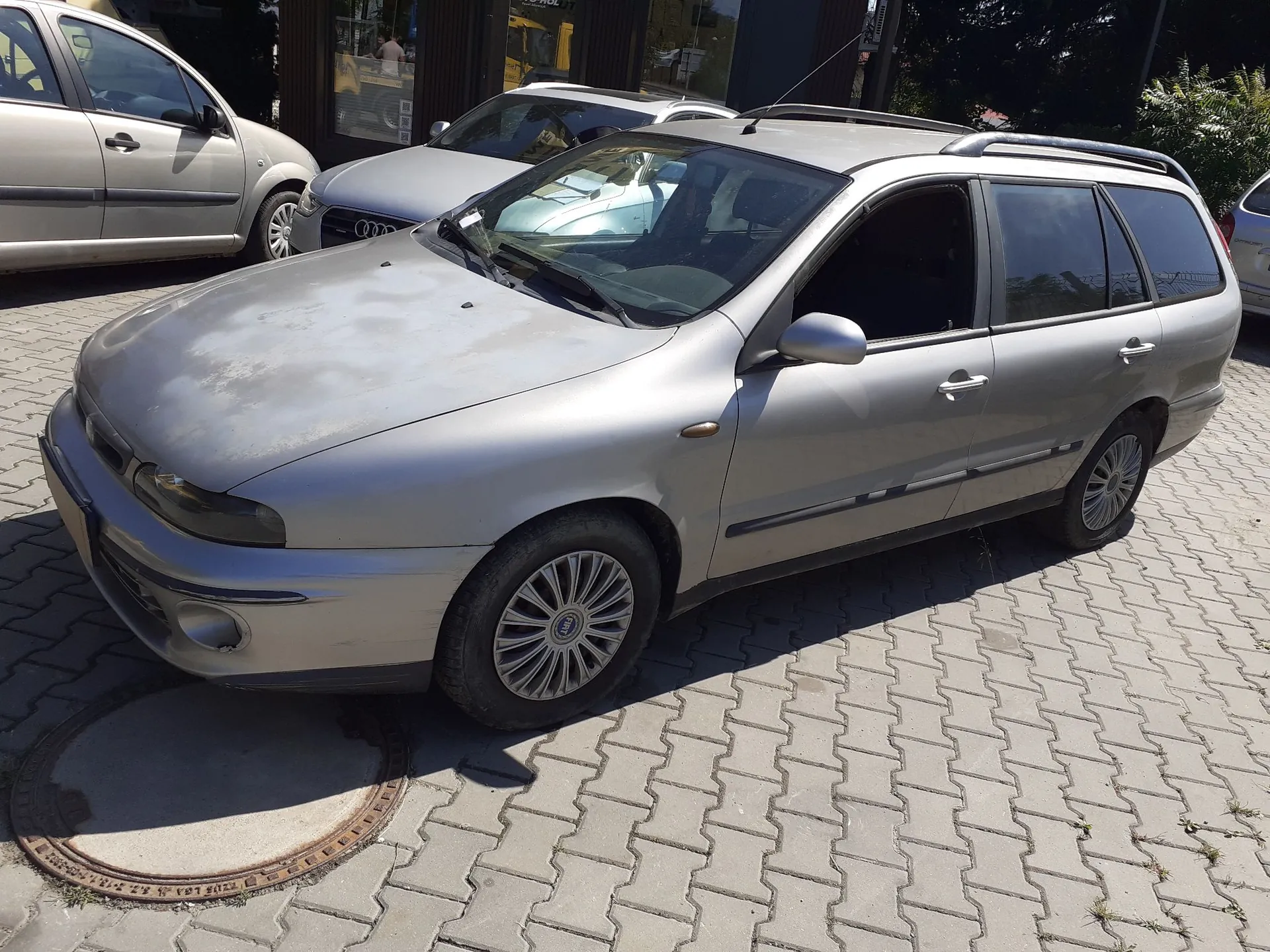 Srebrny samochód typu kombi zaparkowany na kostce brukowej. Pojazd ma widoczne zmatowienia lakieru na masce. W tle widoczne są inne samochody, w tym srebrny Audi oraz jasny samochód osobowy po lewej stronie.