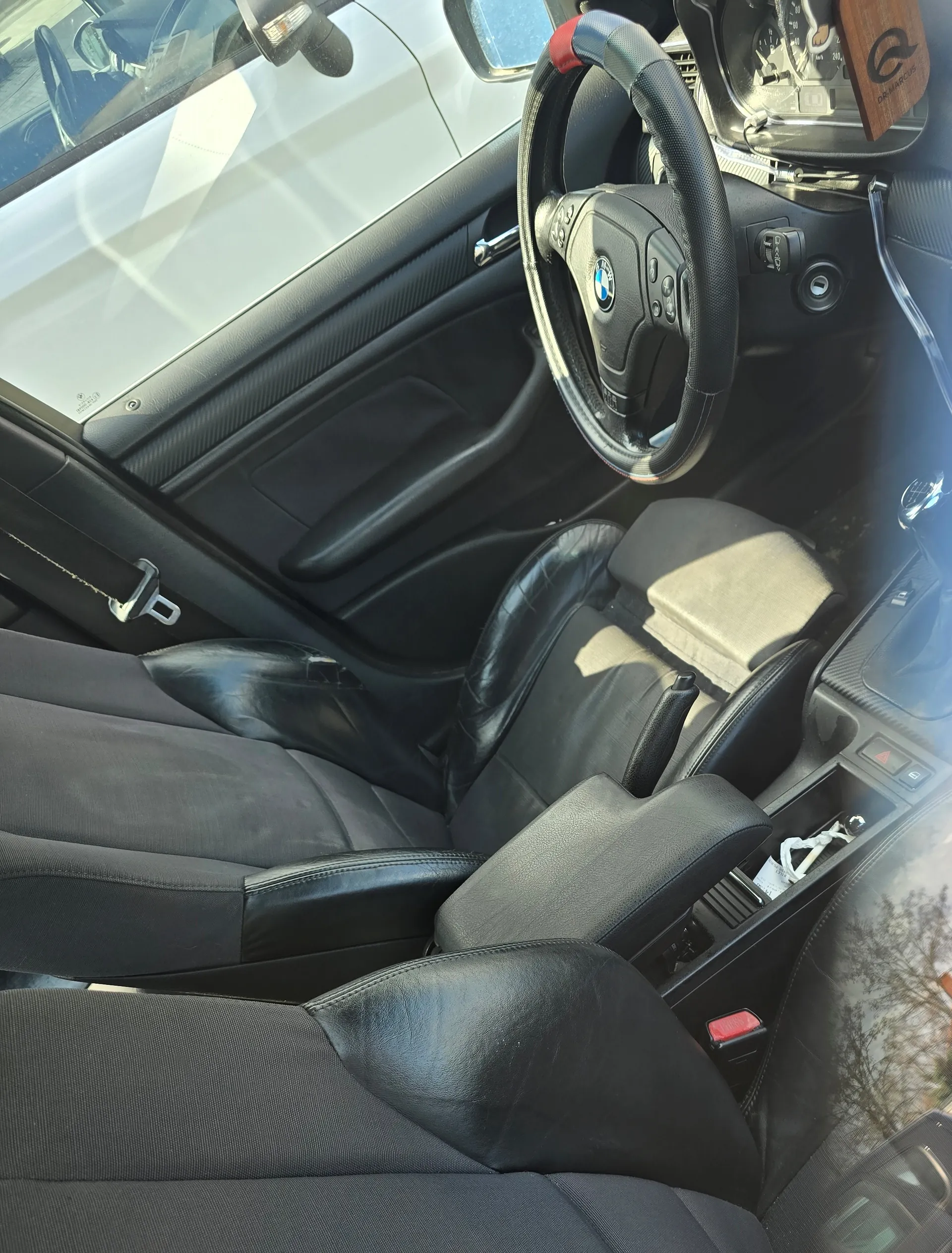 Wnętrze samochodu marki BMW z czarnymi skórzonymi fotelami, kierownicą z logo BMW, dźwignią zmiany biegów i deską rozdzielczą. Widoczna otwarta drzwi pasażera z czarnym panelowaniem i fragmentem białego nadwozia z zewnątrz. Na desce rozdzielczej drewniana tabliczka z logo Q i częściowo widocznym napisem DRIVING. W centralnej konsoli znajduje się przedmiot przypominający białą tasiemkę.