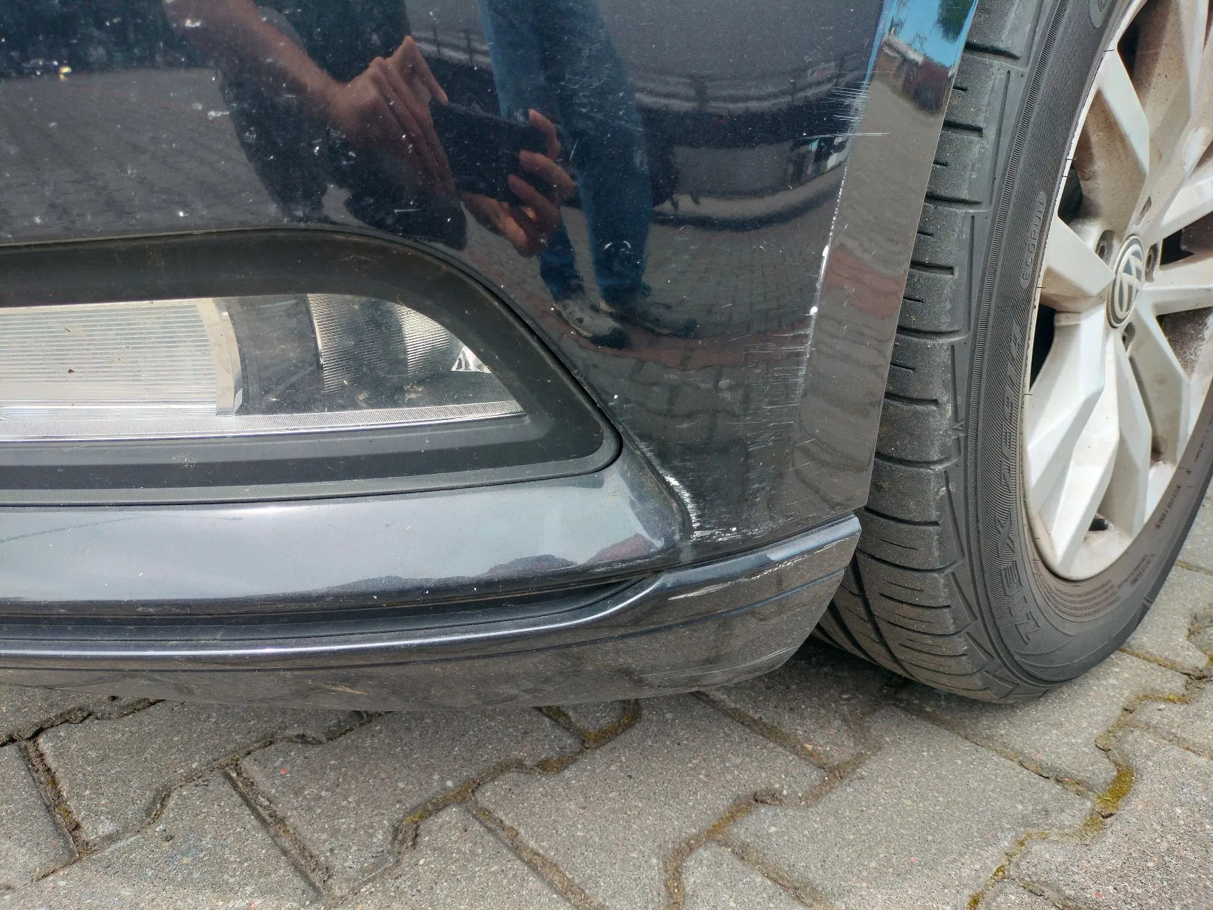 Zbliżenie na przedni prawy róg czarnego samochodu. Na zderzaku widoczne są liczne zarysowania i przetarcia lakieru. Obok zderzaka znajduje się opona z felgą aluminiową. Samochód stoi na kostce brukowej.