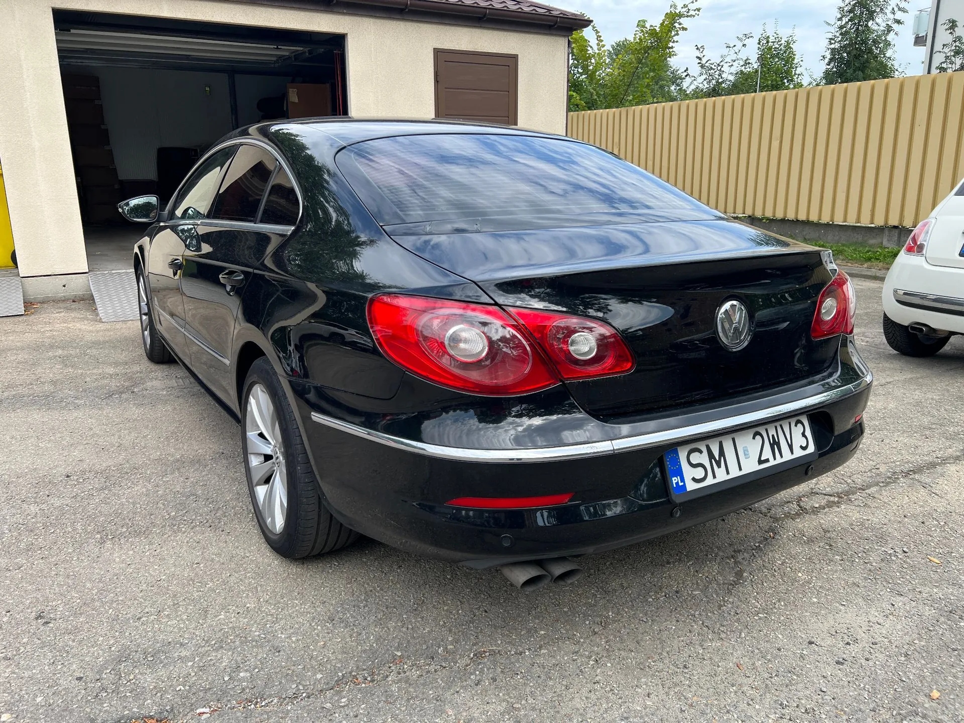 Czarny samochód osobowy marki Volkswagen zaparkowany tyłem na asfaltowym podjeździe przed garażem. Pojazd ma polską tablicę rejestracyjną o numerze SMI 2WV3. W tle widoczny jest biały budynek z otwartym garażem oraz beżowy płot z blachy trapezowej.