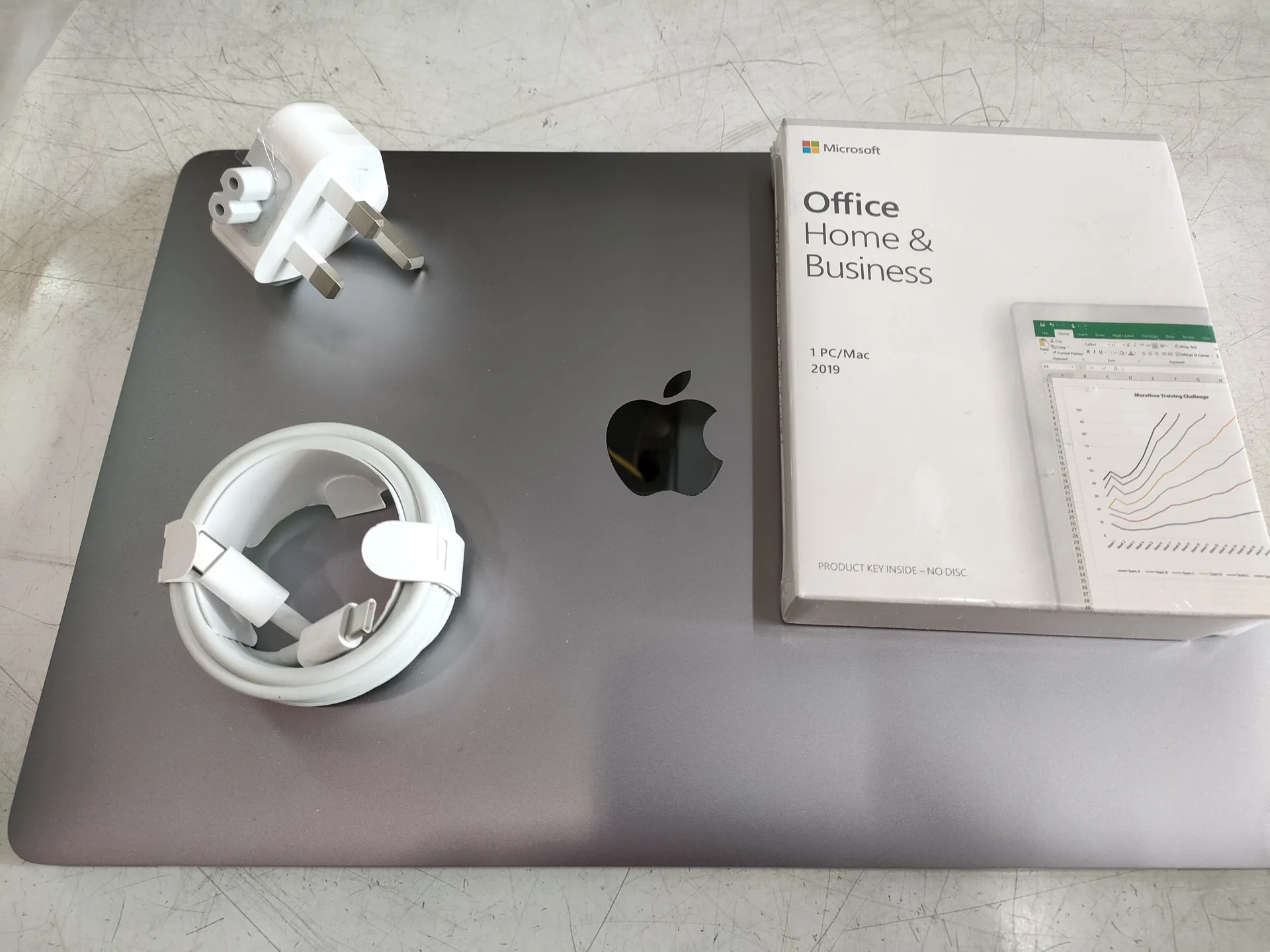 Zamknięty laptop Apple z widocznym logo jabłka na pokrywie. Na laptopie leży biały zasilacz typu UK z trzema szpilkami, zwinięty biały kabel USB-C oraz pudełko Microsoft Office Home & Business 2019. Na pudełku widoczny napis: Microsoft, Office Home & Business, 1 PC/Mac 2019, PRODUCT KEY INSIDE - NO DISC. Po prawej stronie pudełka znajduje się mały obrazek przedstawiający arkusz kalkulacyjny z wykresem liniowym. Tło stanowi metalowa powierzchnia z widocznymi zadrapaniami.