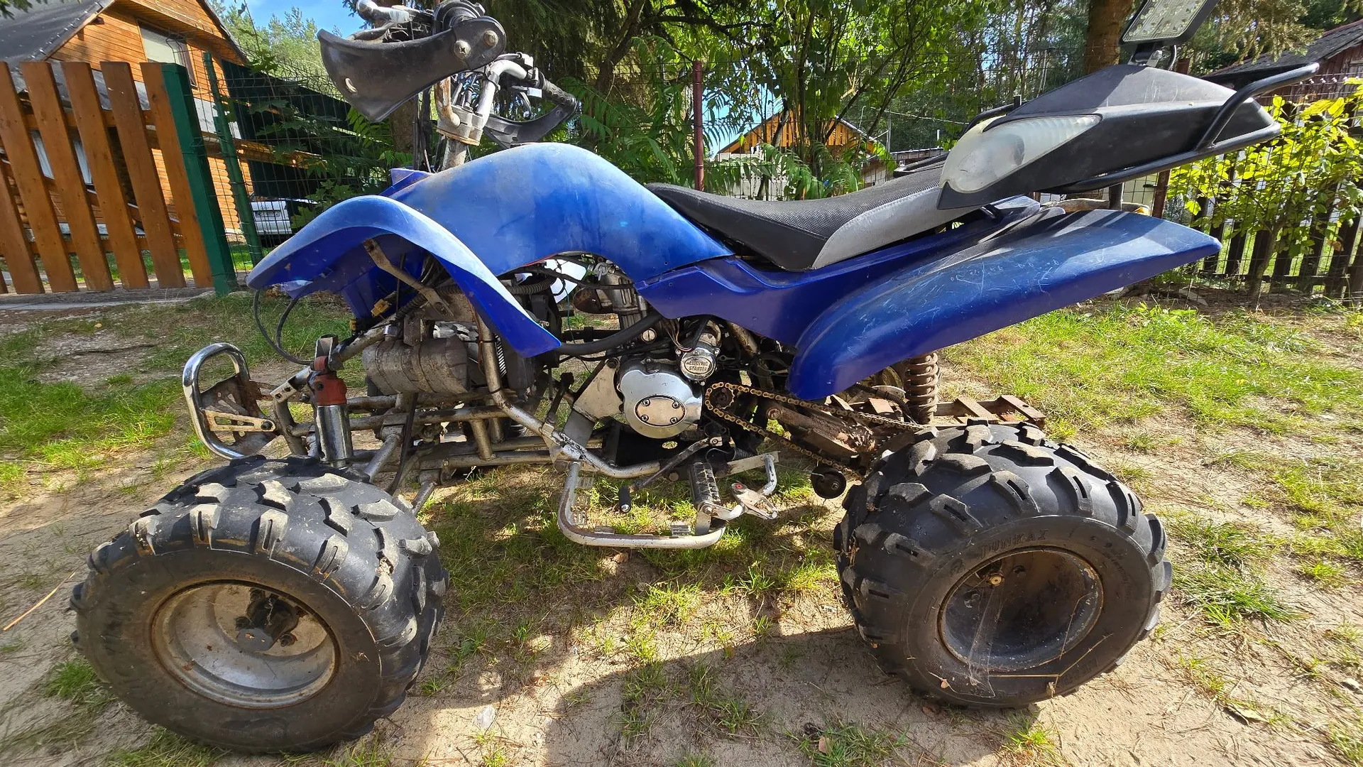 Niebieski quad zaparkowany na trawiasto-piaszczystym terenie. Pojazd ma duże, terenowe opony z wyraźnym bieżnikiem, srebrne ramy ochronne oraz czarne siedzenie. W tle widoczny jest drewniany płot, zielone drzewa i fragmenty budynków.