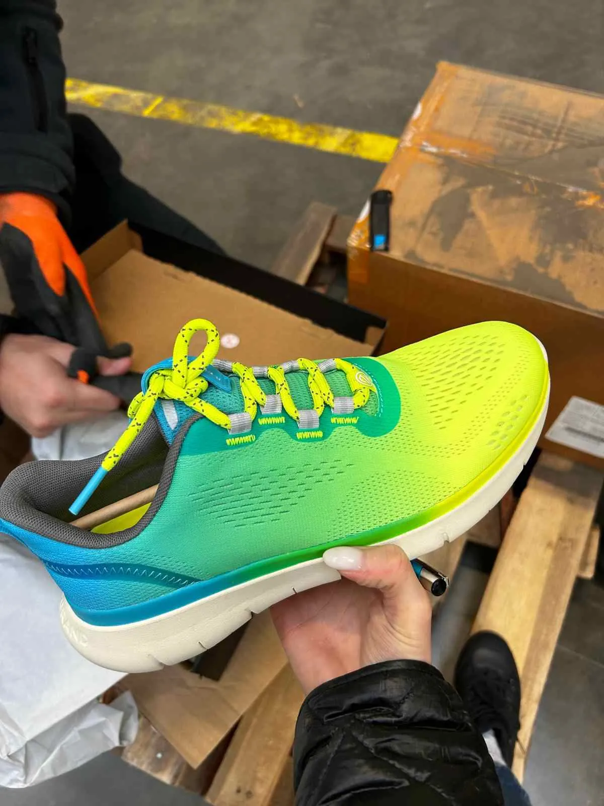 Osoba trzyma w dłoni kolorowy but sportowy z żółtymi sznurówkami, zielono-niebieską górą i białą podeszwą. W tle widoczne są kartony, drewniana paleta, osoba w czarnej kurtce i pomarańczowych rękawiczkach używająca narzędzia do otwierania pudełka oraz żółta linia na betonowej podłodze.