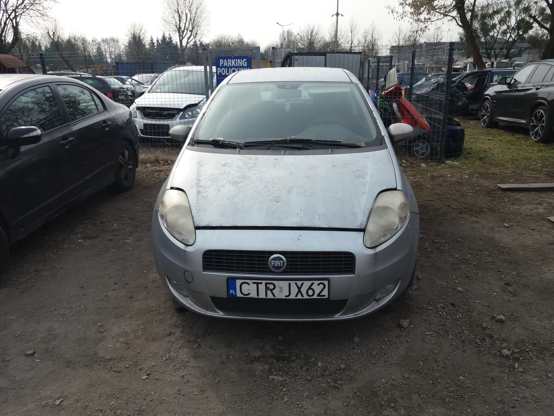 Srebrny samochód marki Fiat z tablicą rejestracyjną CTR JX62 (PL) zaparkowany na brudnej, żwirowej powierzchni. W tle widoczne są inne pojazdy, w tym uszkodzony czerwony samochód za ogrodzeniem, czarny samochód po lewej stronie oraz biały samochód za ogrodzeniem. Na tle niebieski znak z napisem PARKING POLICY. Tło stanowią drzewa bez liści, ogrodzenie i pochmurne niebo.